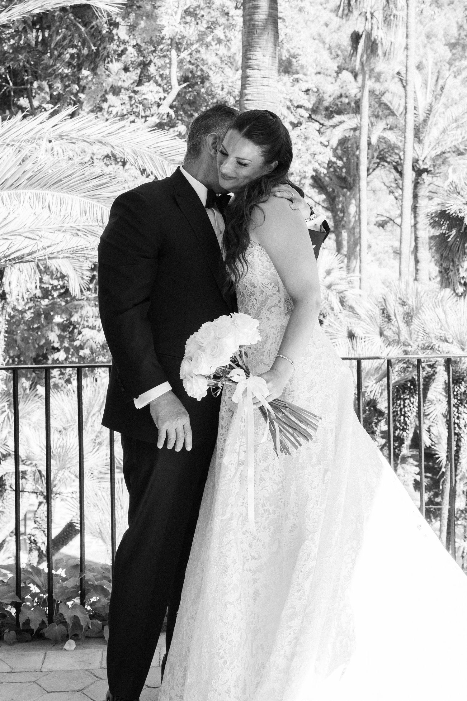 Black and white photo of a bride and groom sharing a romantic kiss on a balcony surrounded by lush tropical greenery.