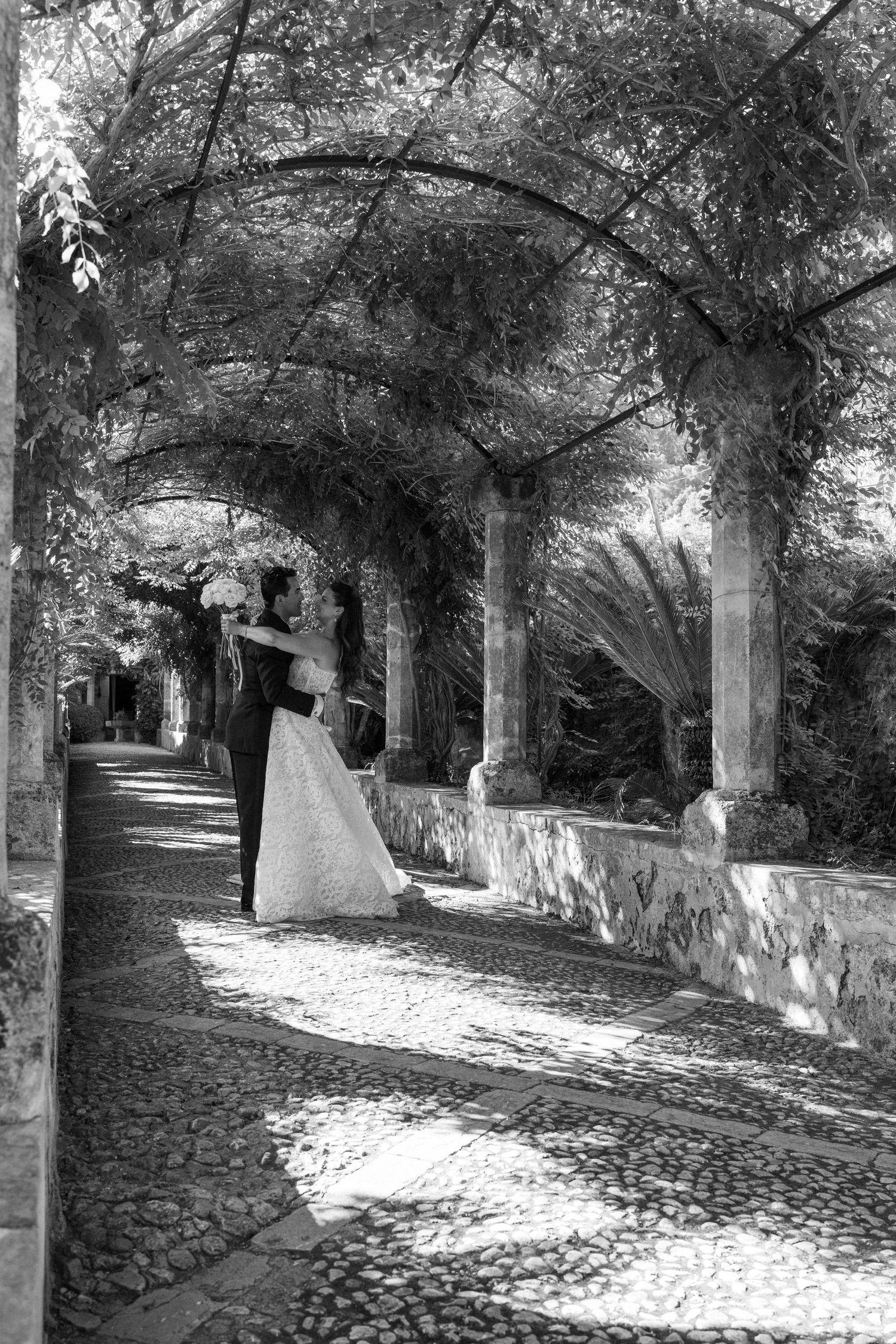 Black and white photo of a bride and groom embracing under a vine-covered stone archway on a cobblestone path.