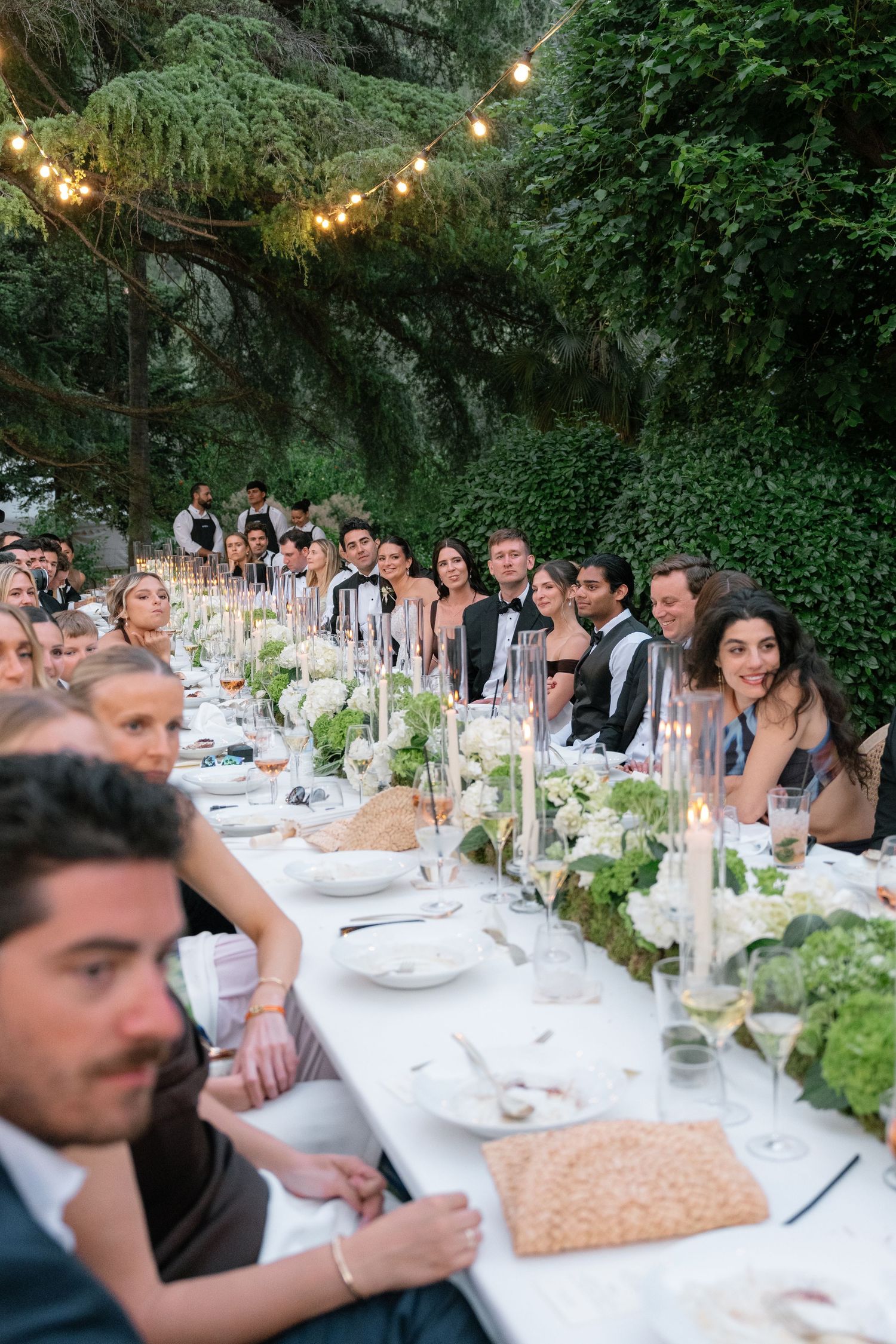 Elegant outdoor wedding reception with guests seated at a long table adorned with white flowers, candles, and string lights.