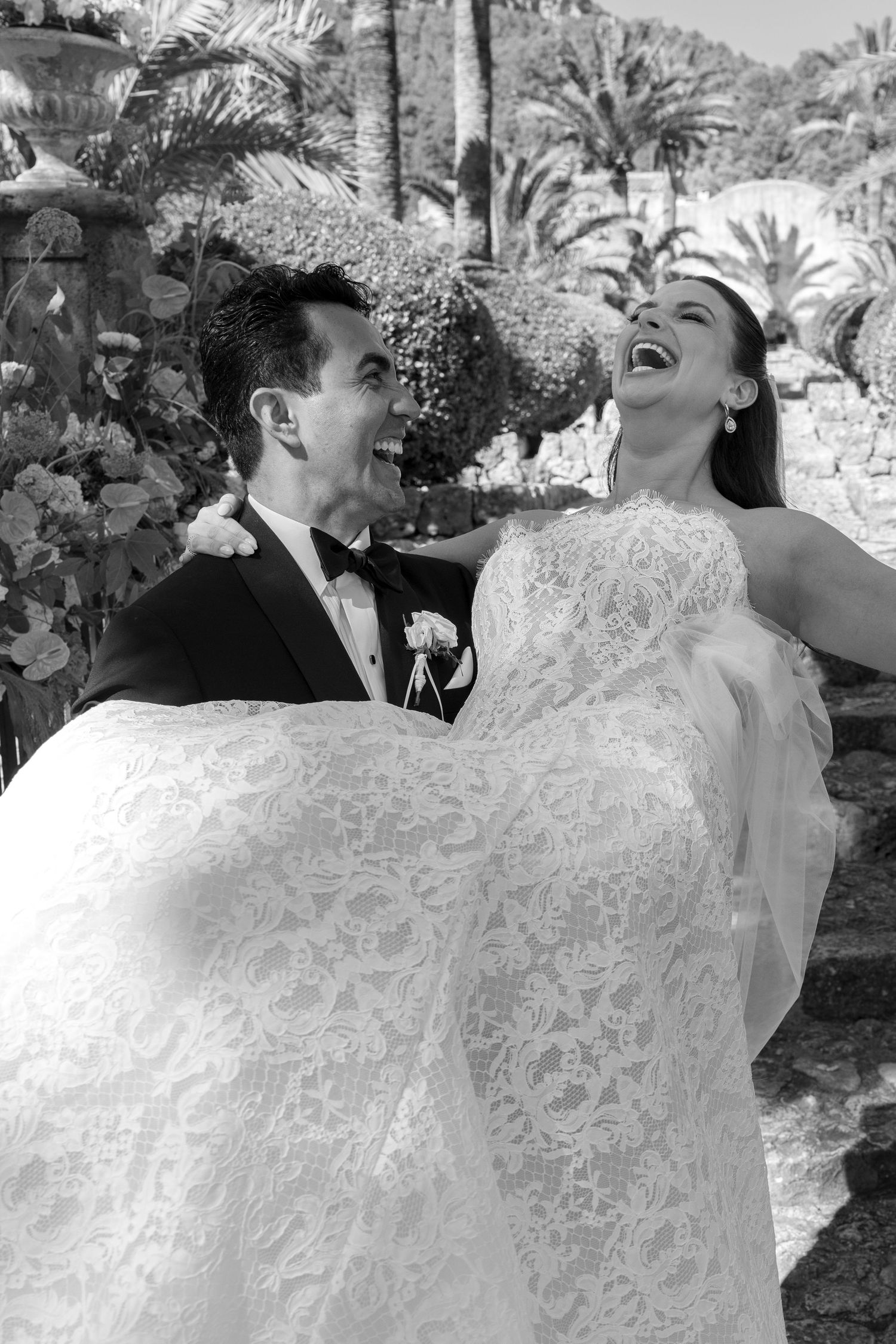 Black and white photo of a joyful bride and groom laughing together outdoors, surrounded by palm trees on their wedding day.
