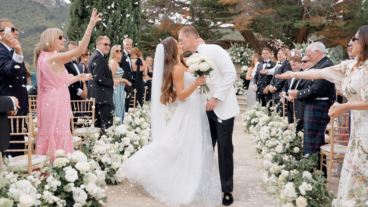 Newlywed couple sharing a kiss at outdoor wedding ceremony while guests toss confetti along floral aisle.