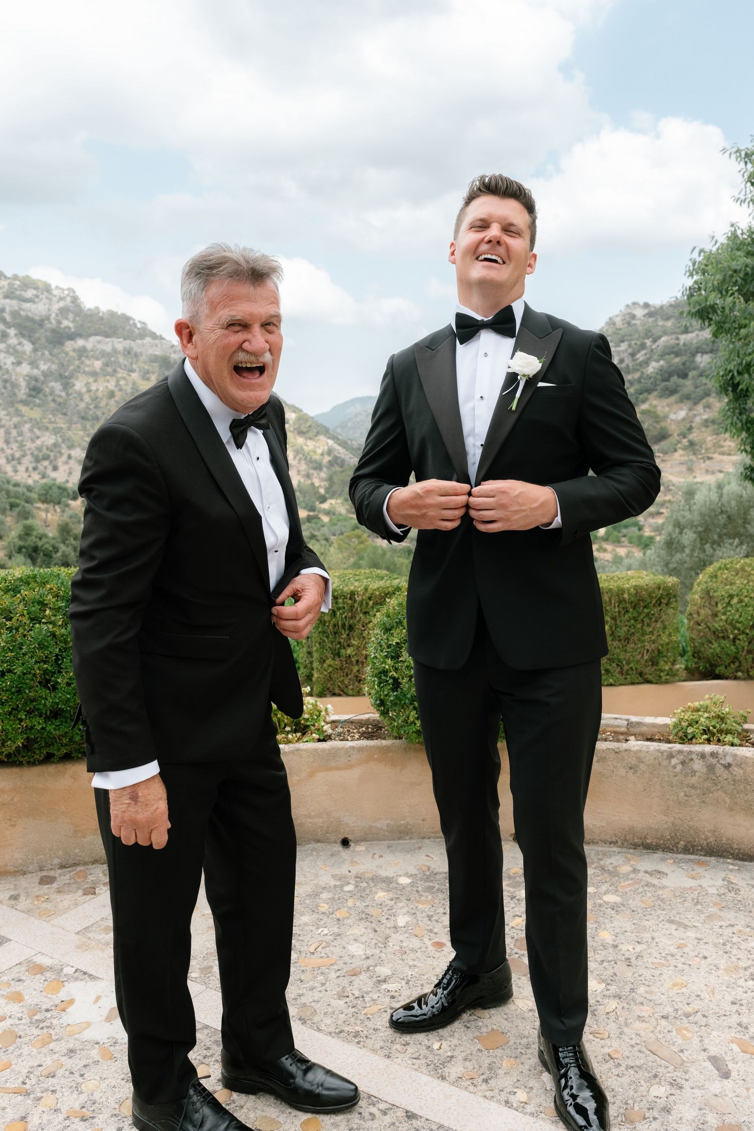 Two men in black tuxedos laughing joyfully outdoors with mountain scenery, likely a groom and father at a wedding.