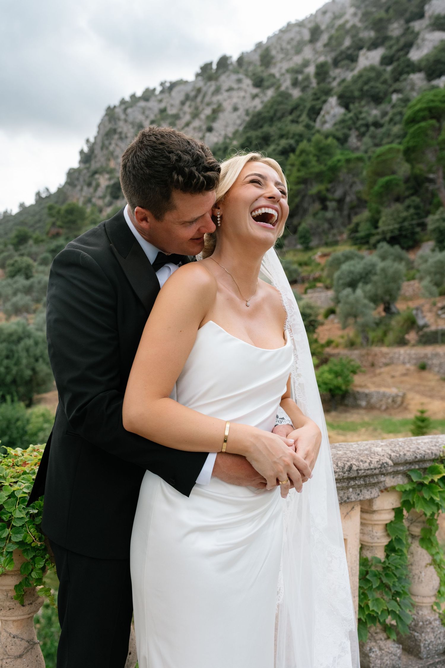 Bride in white gown laughing joyfully as groom in black tuxedo embraces her on a stone terrace with mountain backdrop.