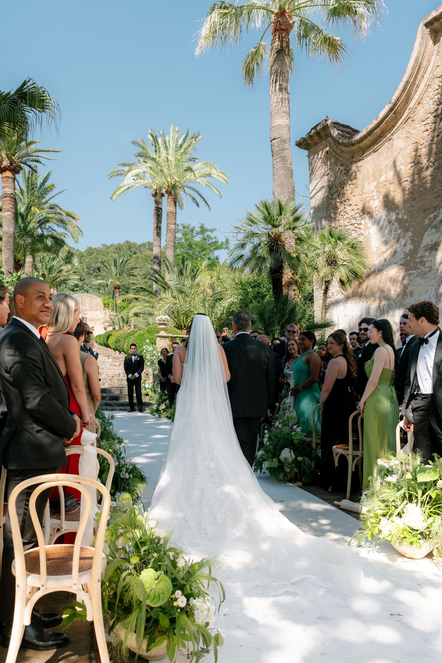 Outdoor wedding ceremony with bride in veil walking the aisle, palm trees and stone ruins backdrop.
