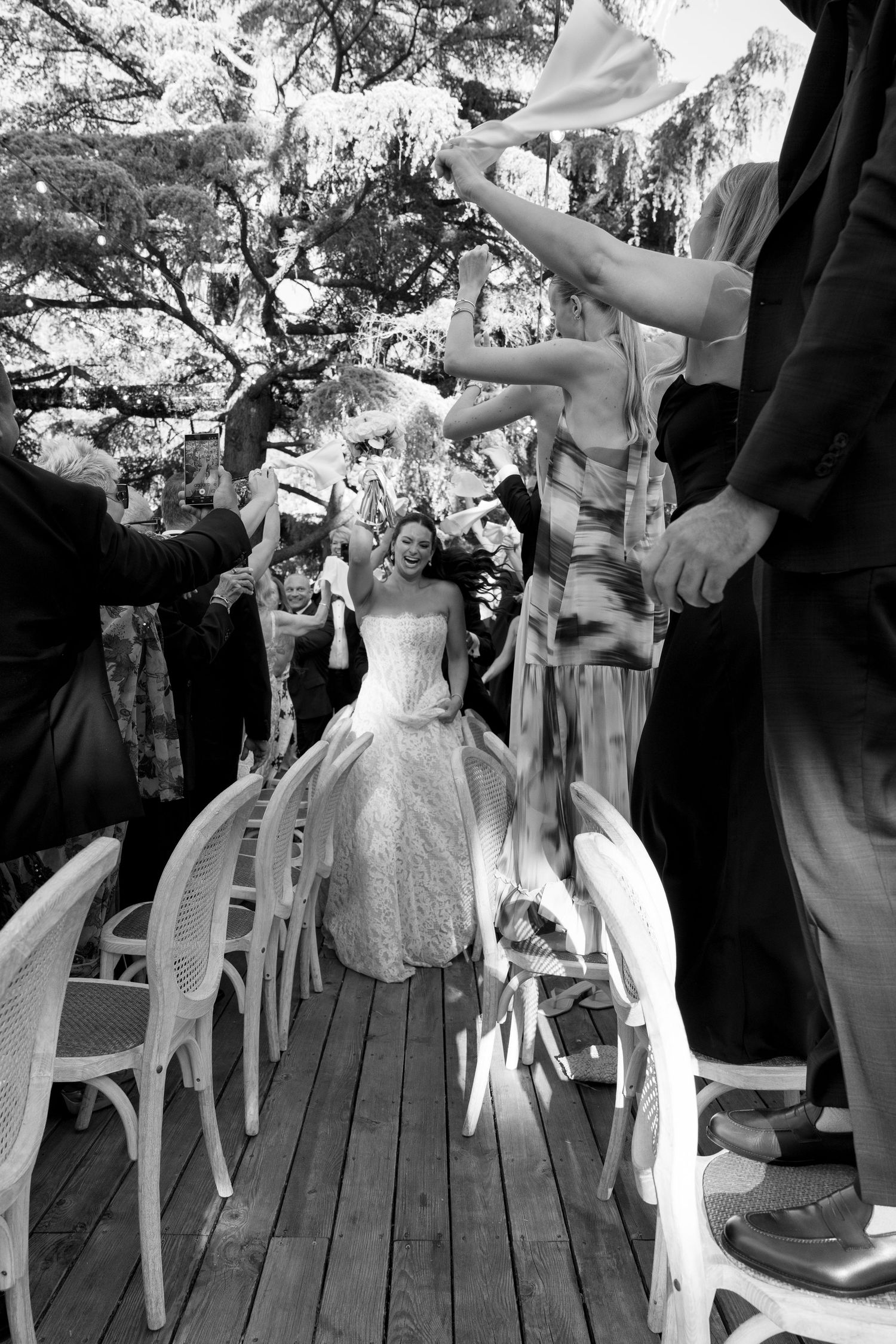 Black and white photo of a smiling bride walking down the aisle at an outdoor wedding ceremony.