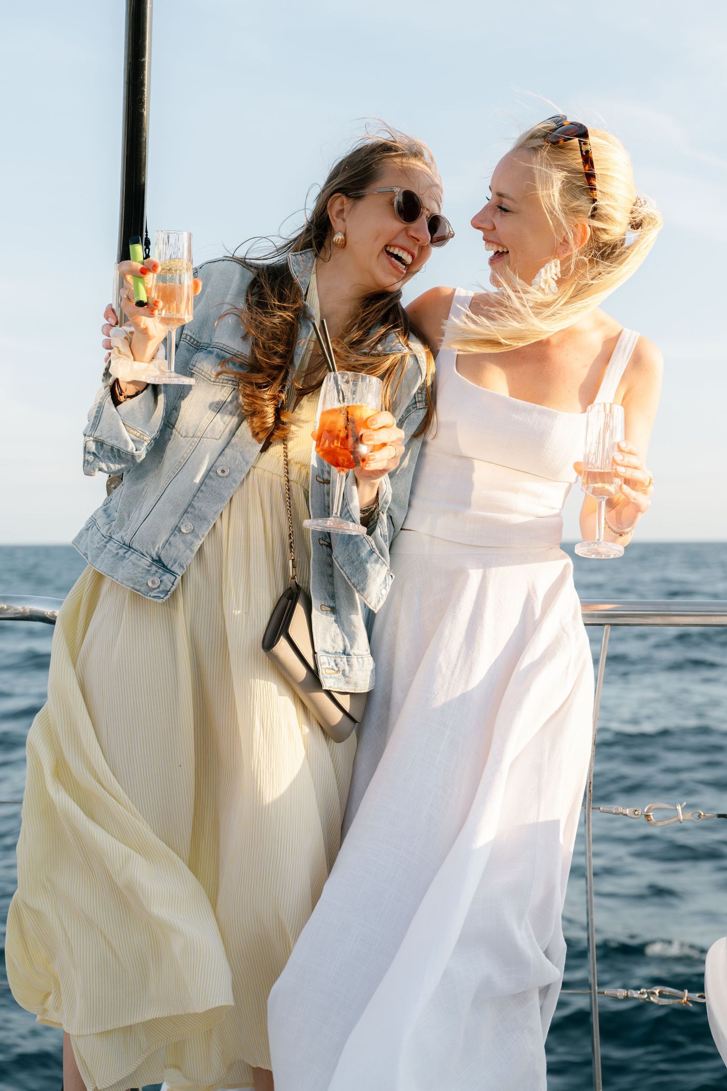 Two friends in light colored dresses toast with drinks against an ocean backdrop.