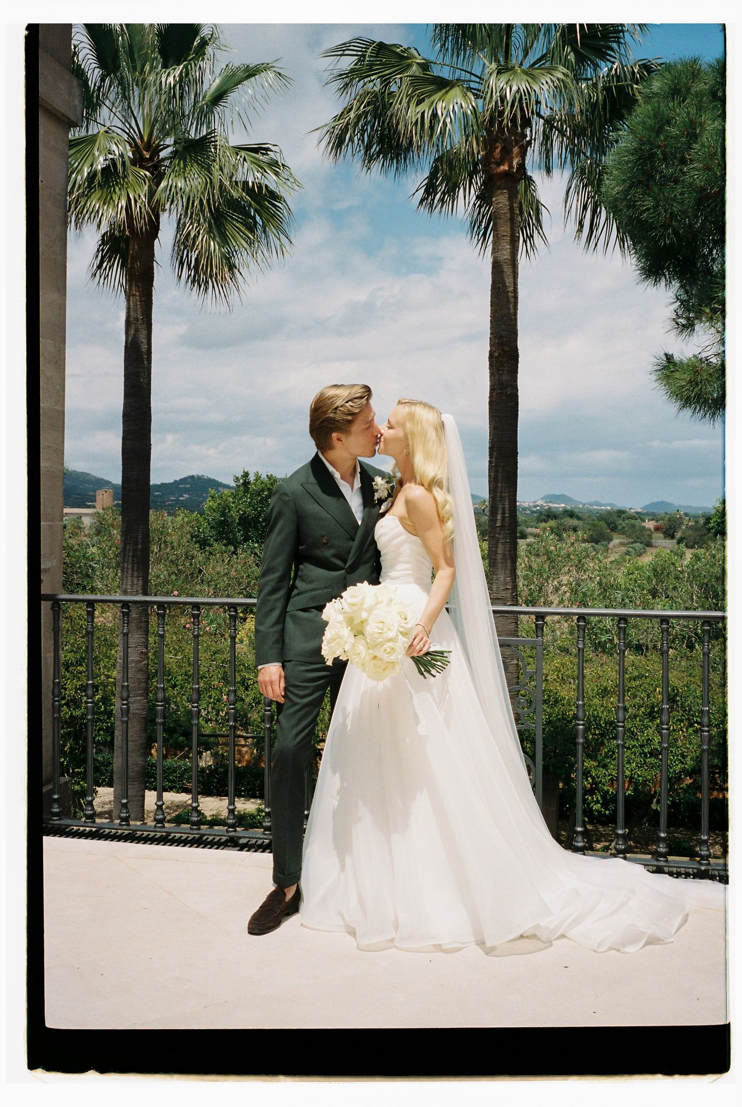 Wedding couple sharing an intimate moment on a terrace with palm trees and mountain views.