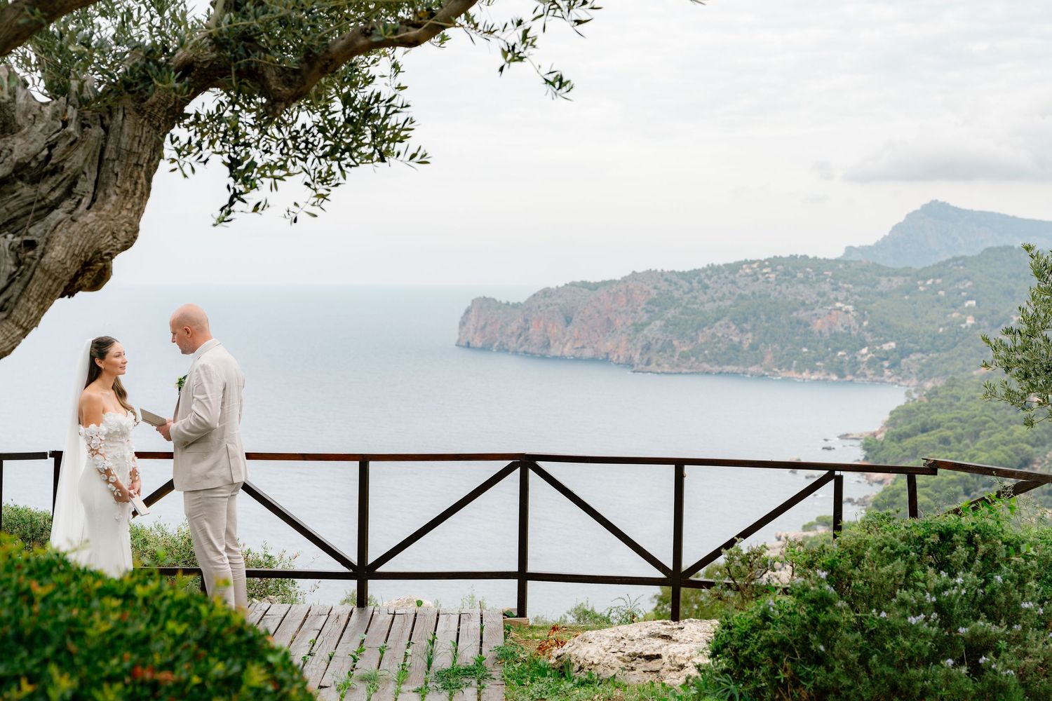 Scenic Mediterranean coastline view with mountains and wooden deck railing in foreground.