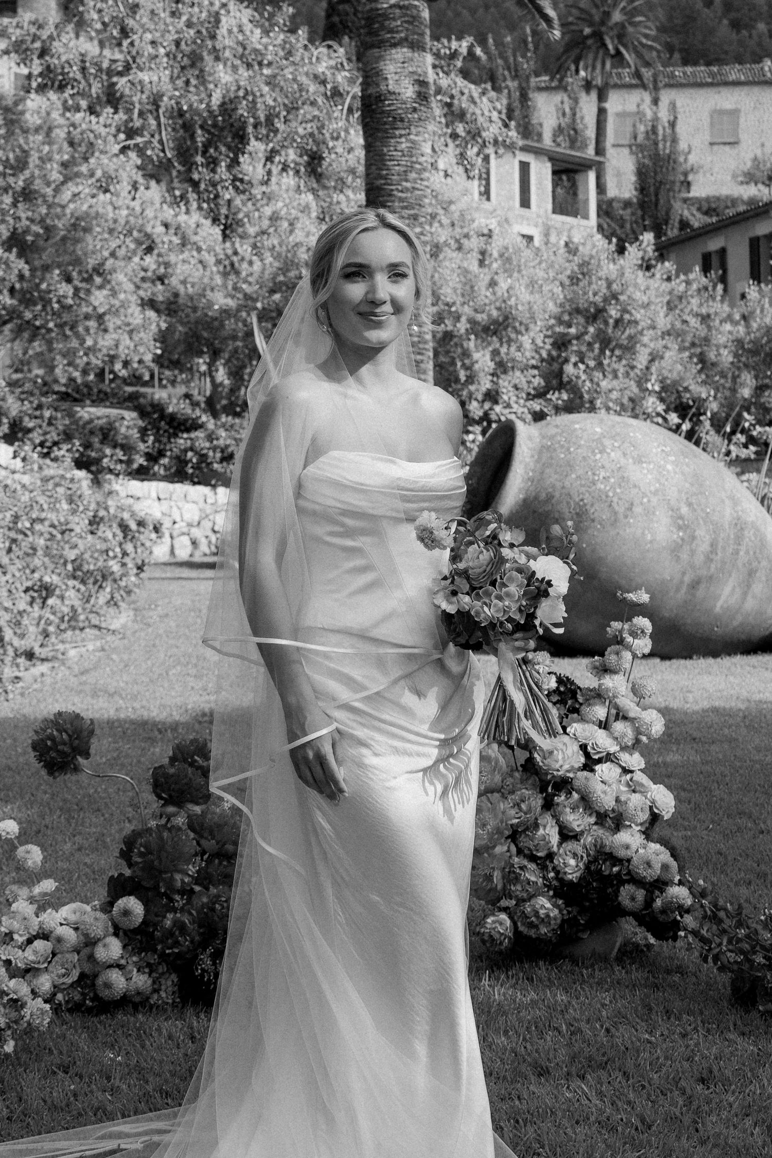 Elegant bride in white dress posing in a garden with blooming flowers.