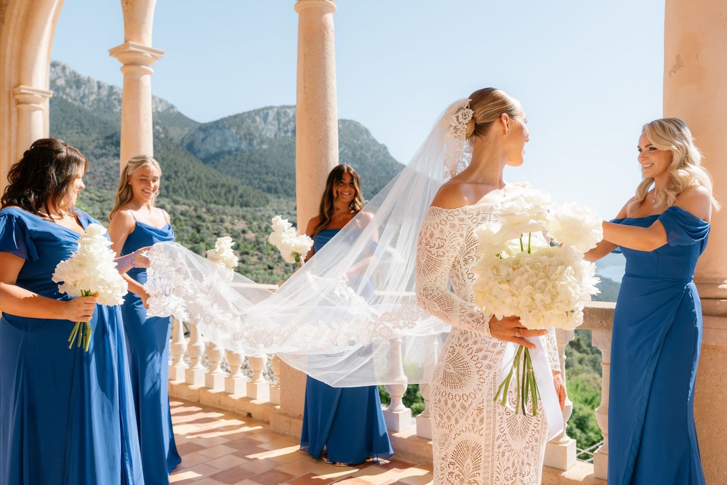 Wedding party in royal blue dresses helping with the bride's veil on a scenic terrace.