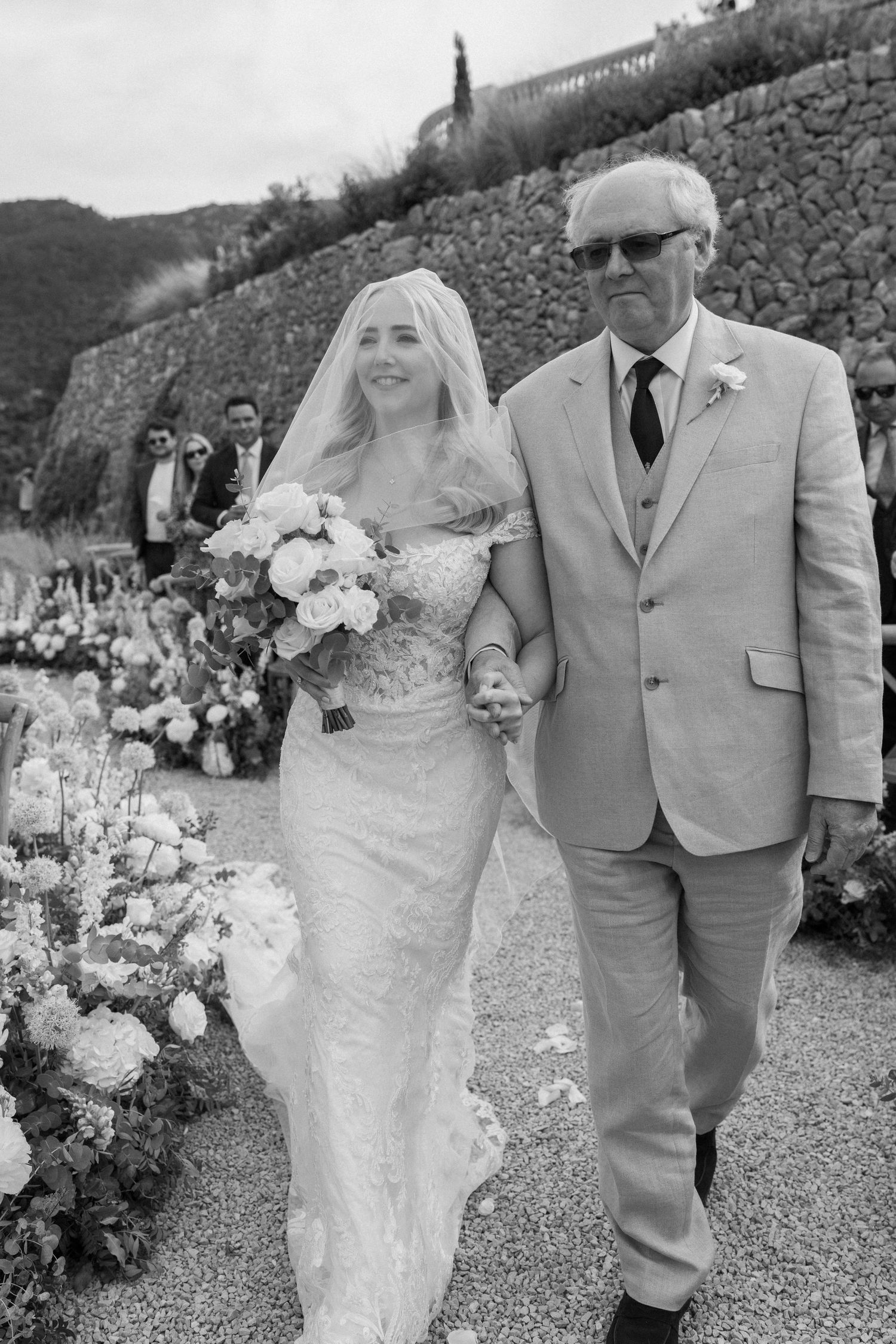A wedding processional along a flower-lined path with mountain scenery.