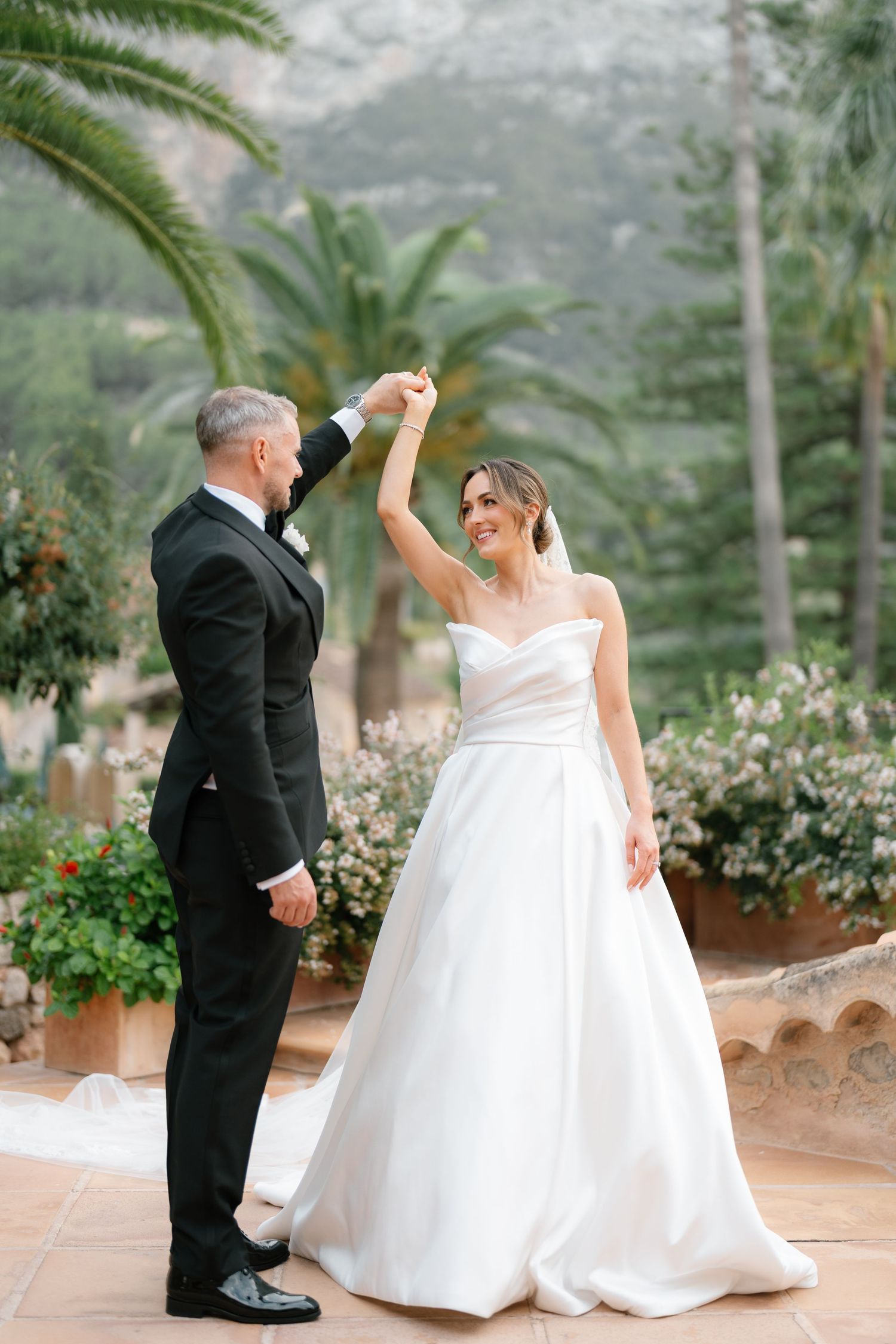 A couple in wedding attire dances together on an outdoor patio surrounded by lush greenery.