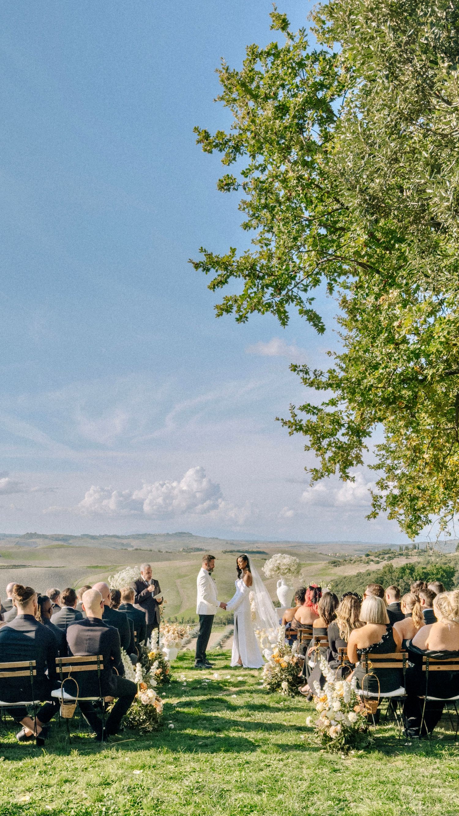 A scenic outdoor wedding ceremony set against rolling hills with guests seated on wooden benches under a blue sky.
