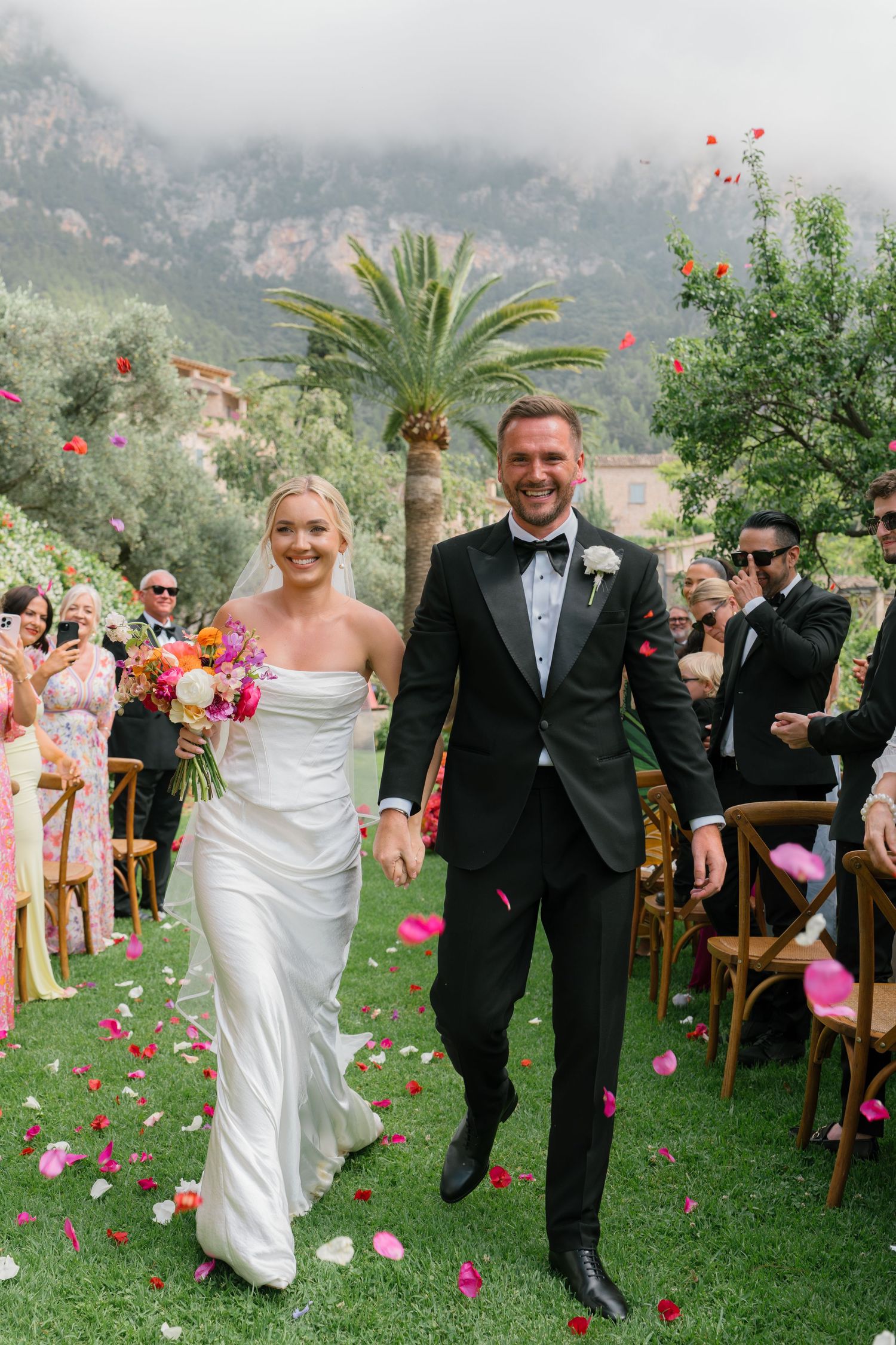 Happy couple walks down aisle surrounded by pink flower petals and mountain scenery.
