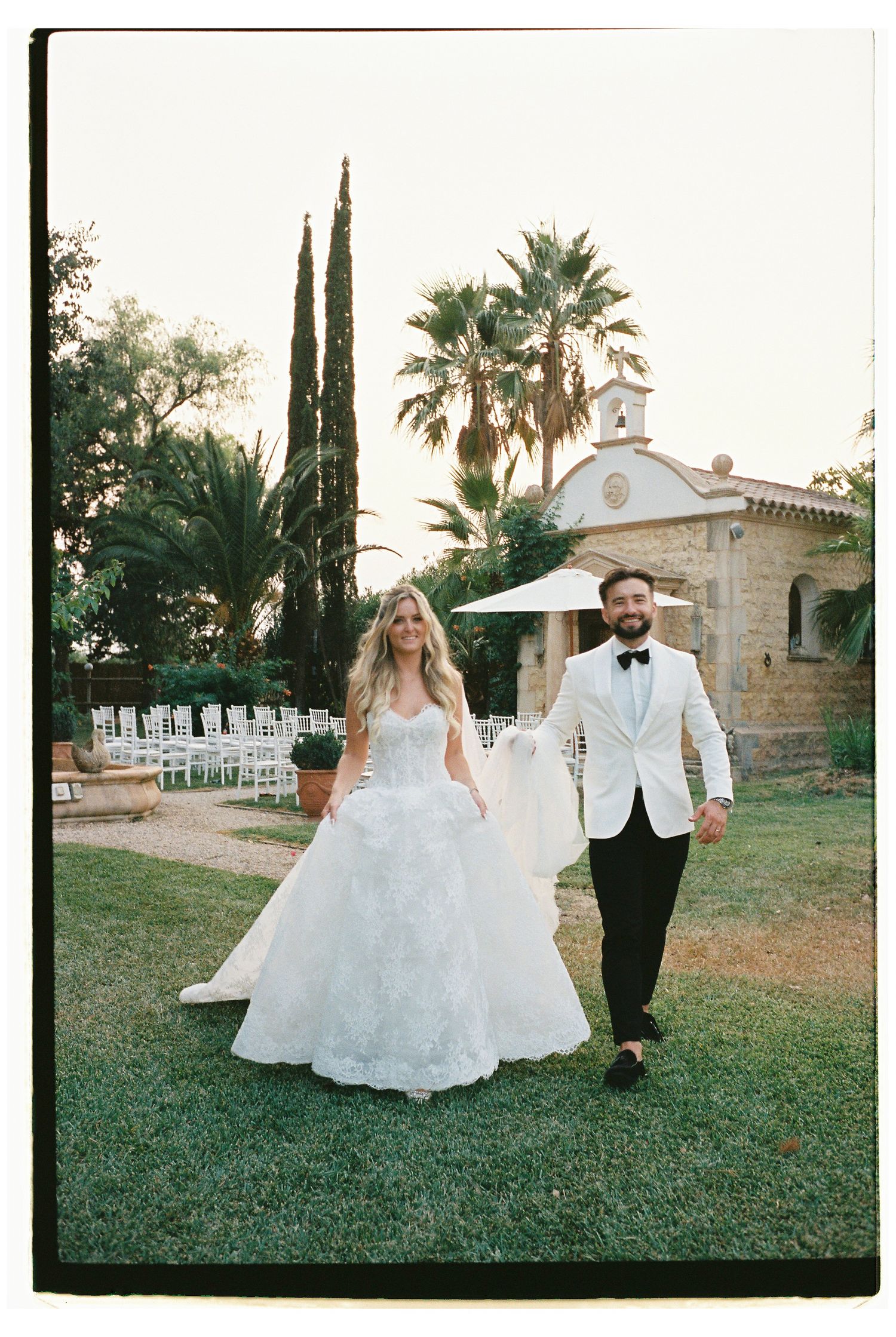 Newlyweds walking hand in hand on the lawn surrounded by palm trees and a stone building at Finca Santa Lucia, Mallorca