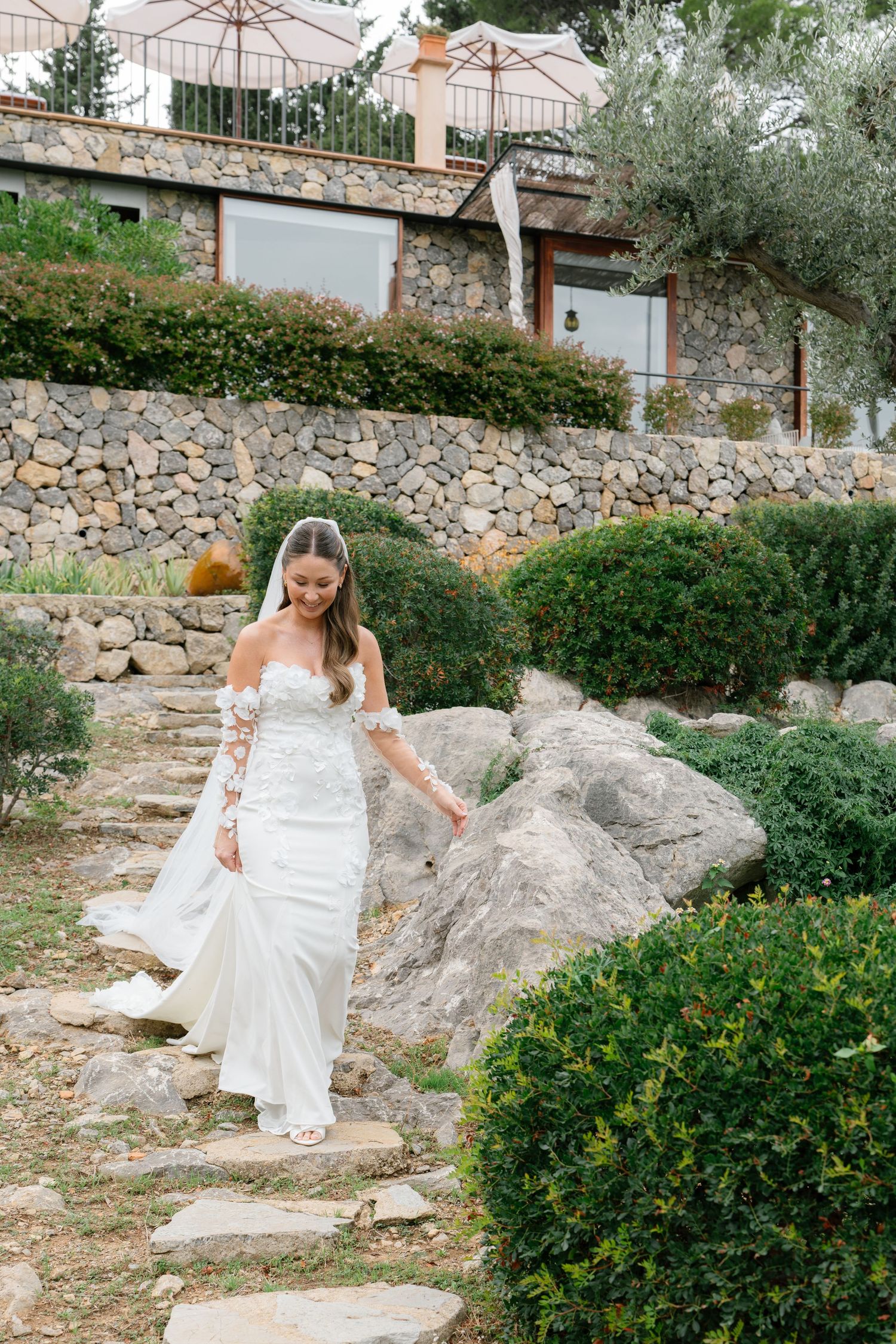 Bride in white dress walking through Mediterranean garden with stone walls and olive trees.