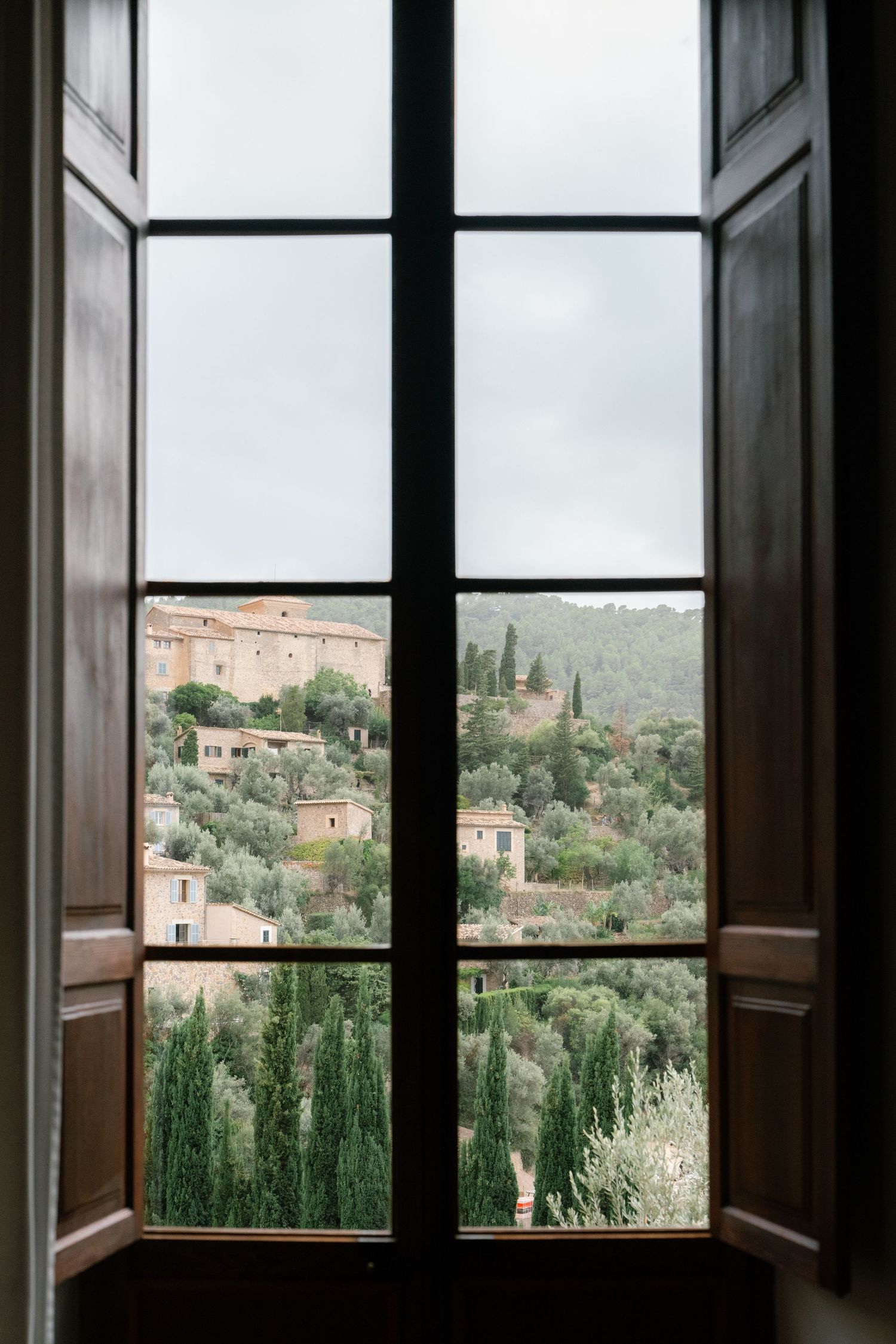 View through an open window overlooking a Mediterranean hillside village with cypress trees and stone buildings.