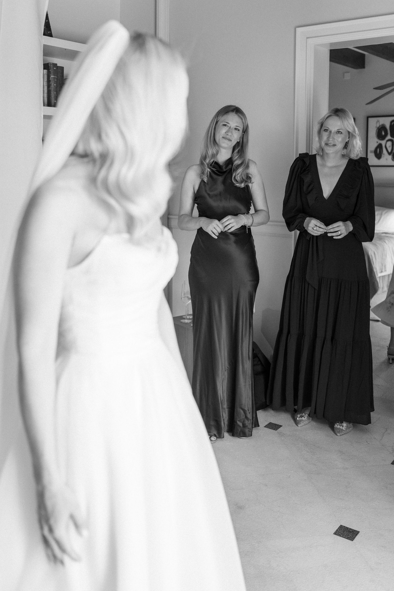 Wedding preparation scene with bridesmaids in black dresses watching bride in white gown.