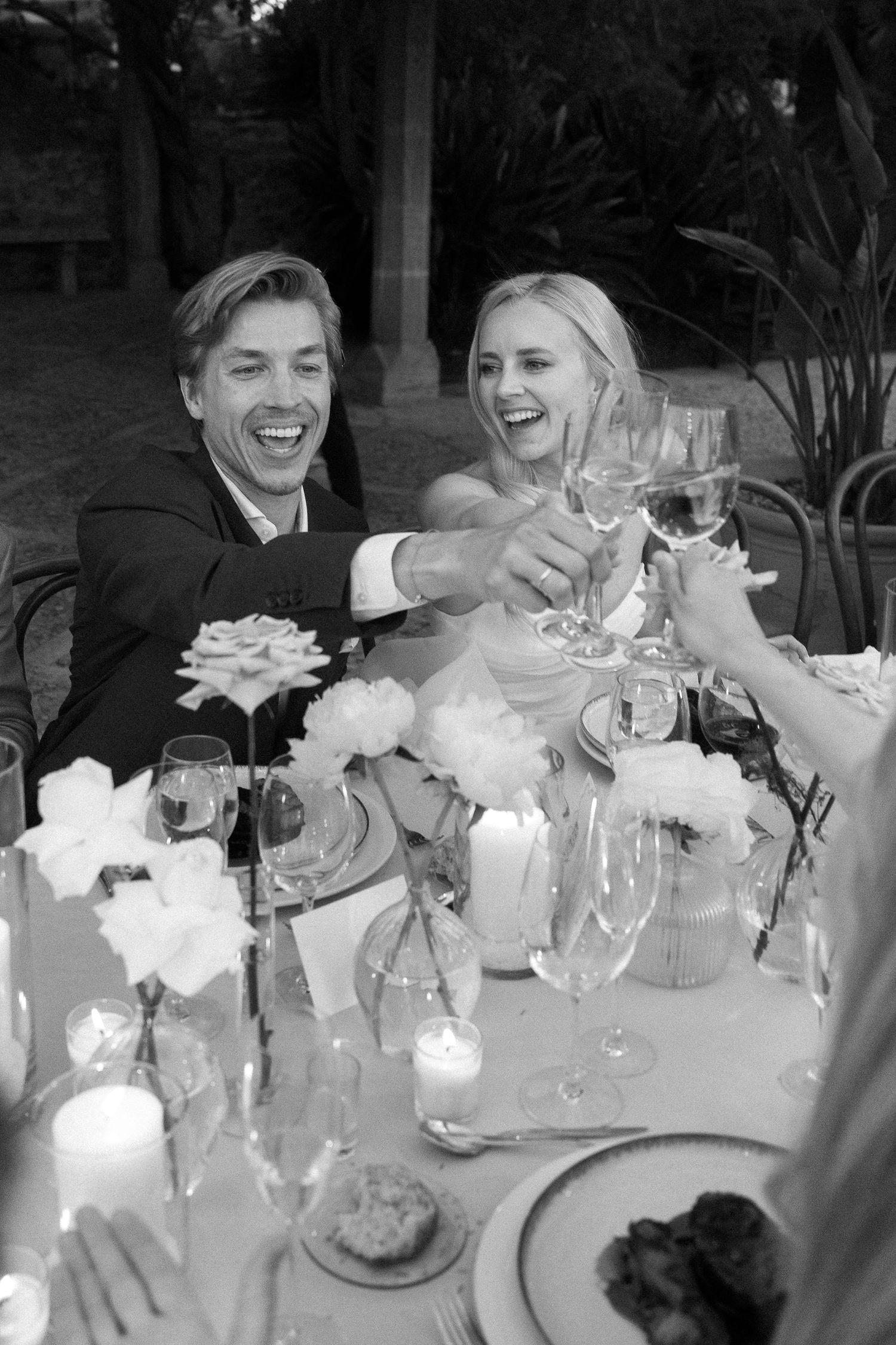 Couple laughing while toasting at an elegant outdoor wedding reception with floral centerpieces.