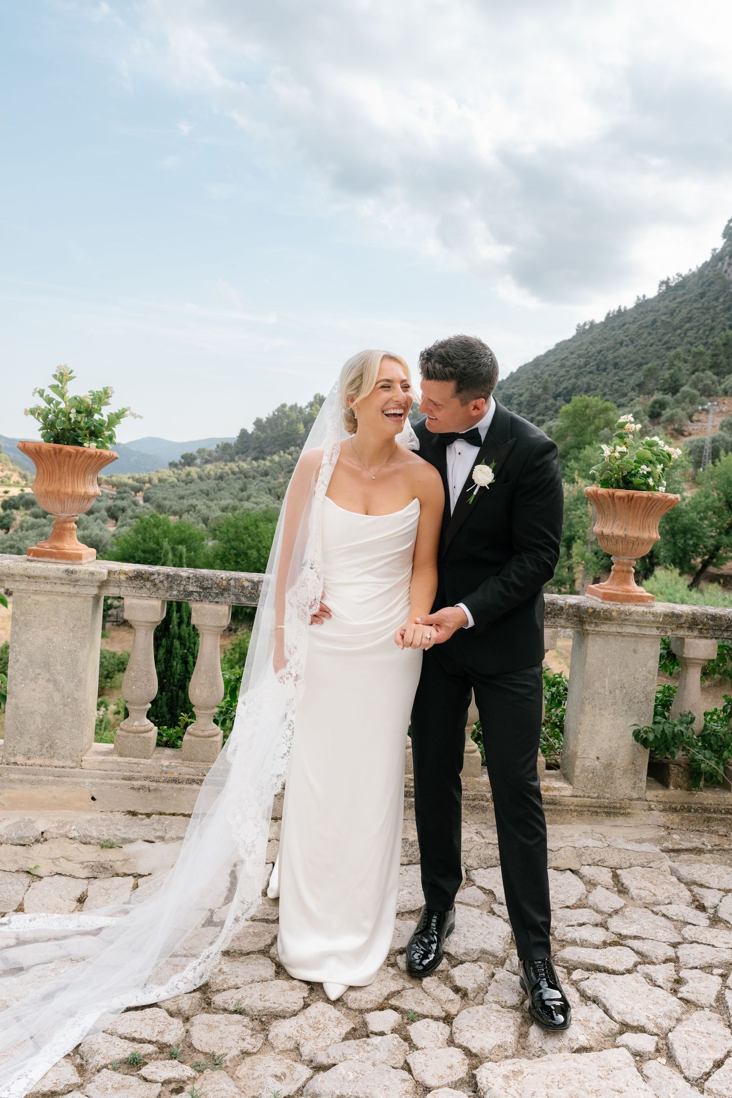Bride in white gown and veil laughs as groom in black tuxedo whispers to her on a stone terrace with lush hills.