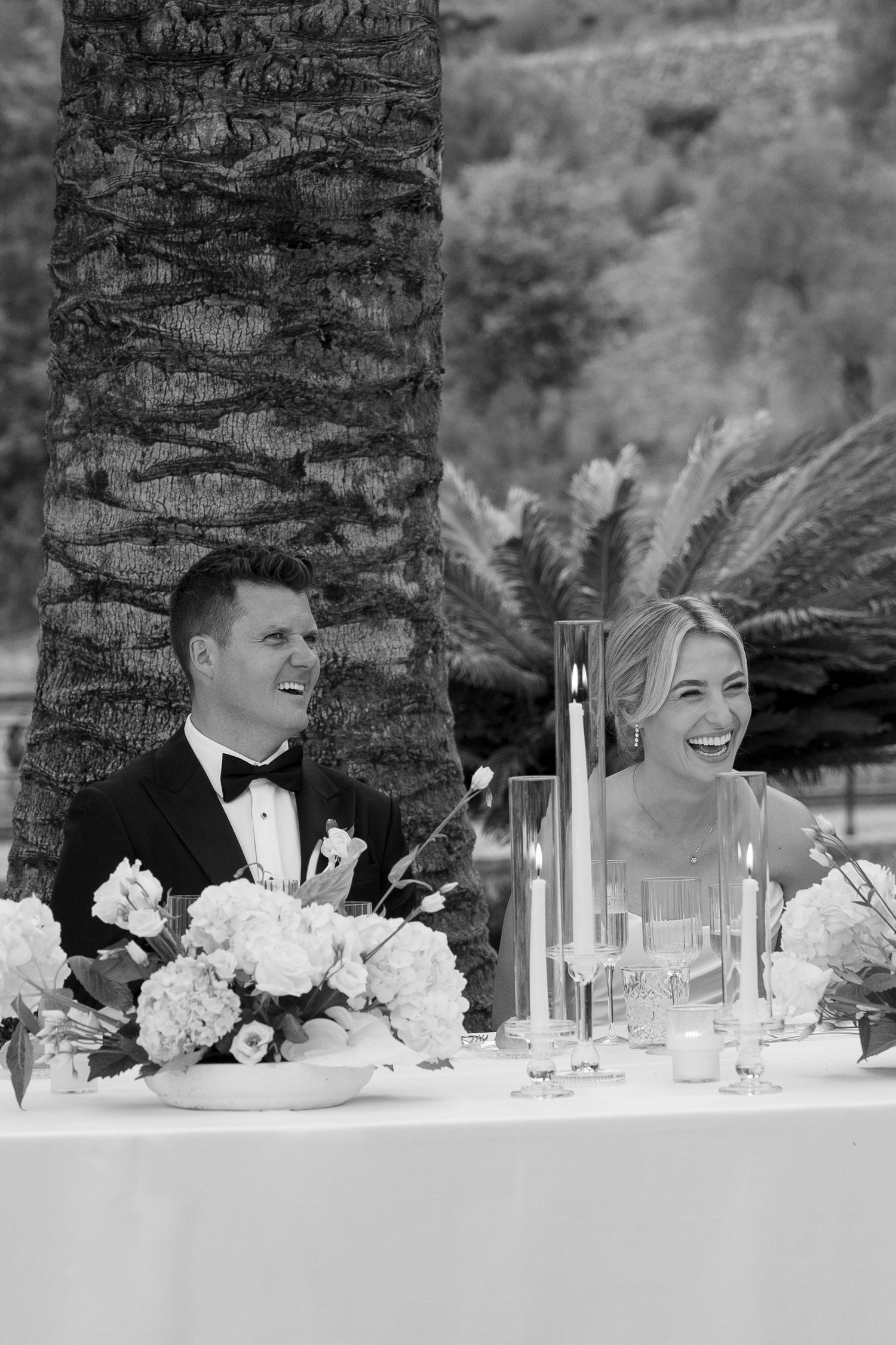 Black and white photo of a laughing bride and groom seated at their wedding reception table with floral arrangements.