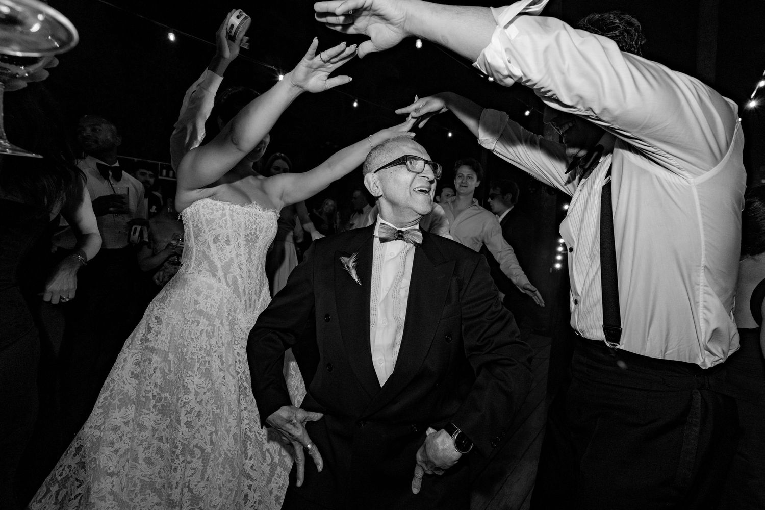Black and white wedding reception dance floor photo of elderly man in tuxedo dancing joyfully surrounded by guests.
