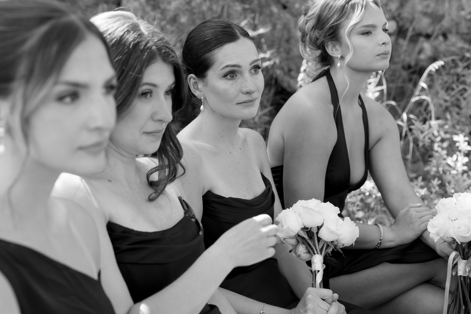 Four bridesmaids in black dresses sitting together outdoors, holding white flowers, in black and white photography.