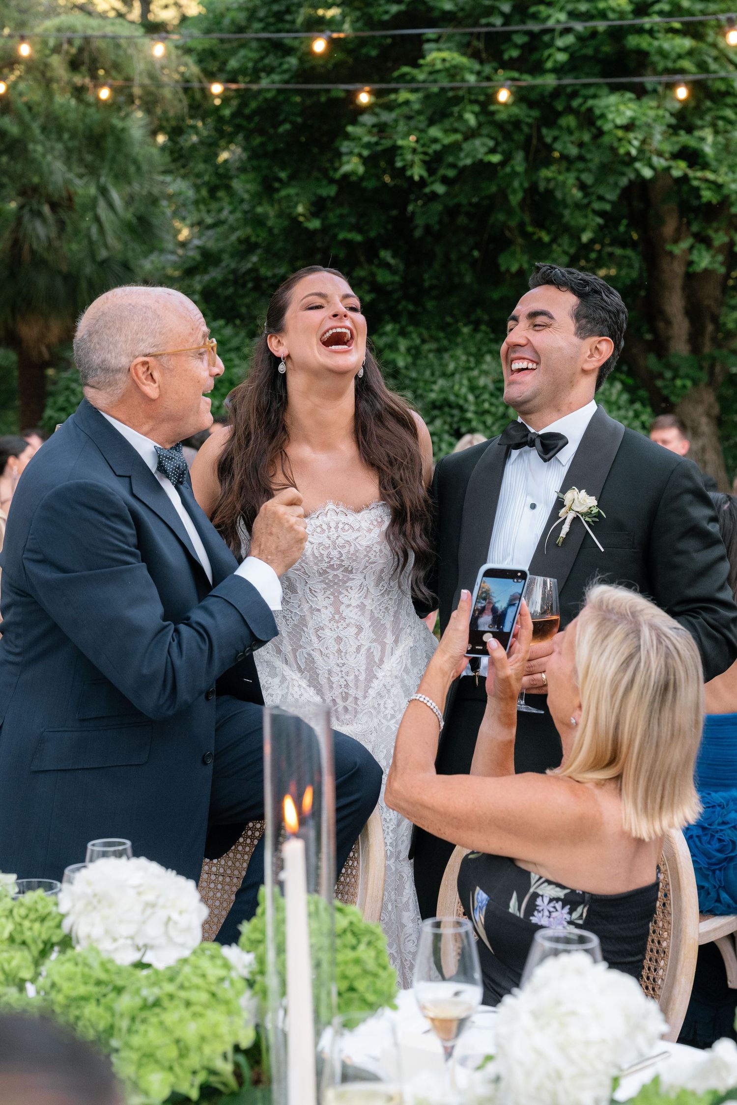 Bride and groom laughing joyfully with older man at outdoor wedding reception while guest photographs the moment.