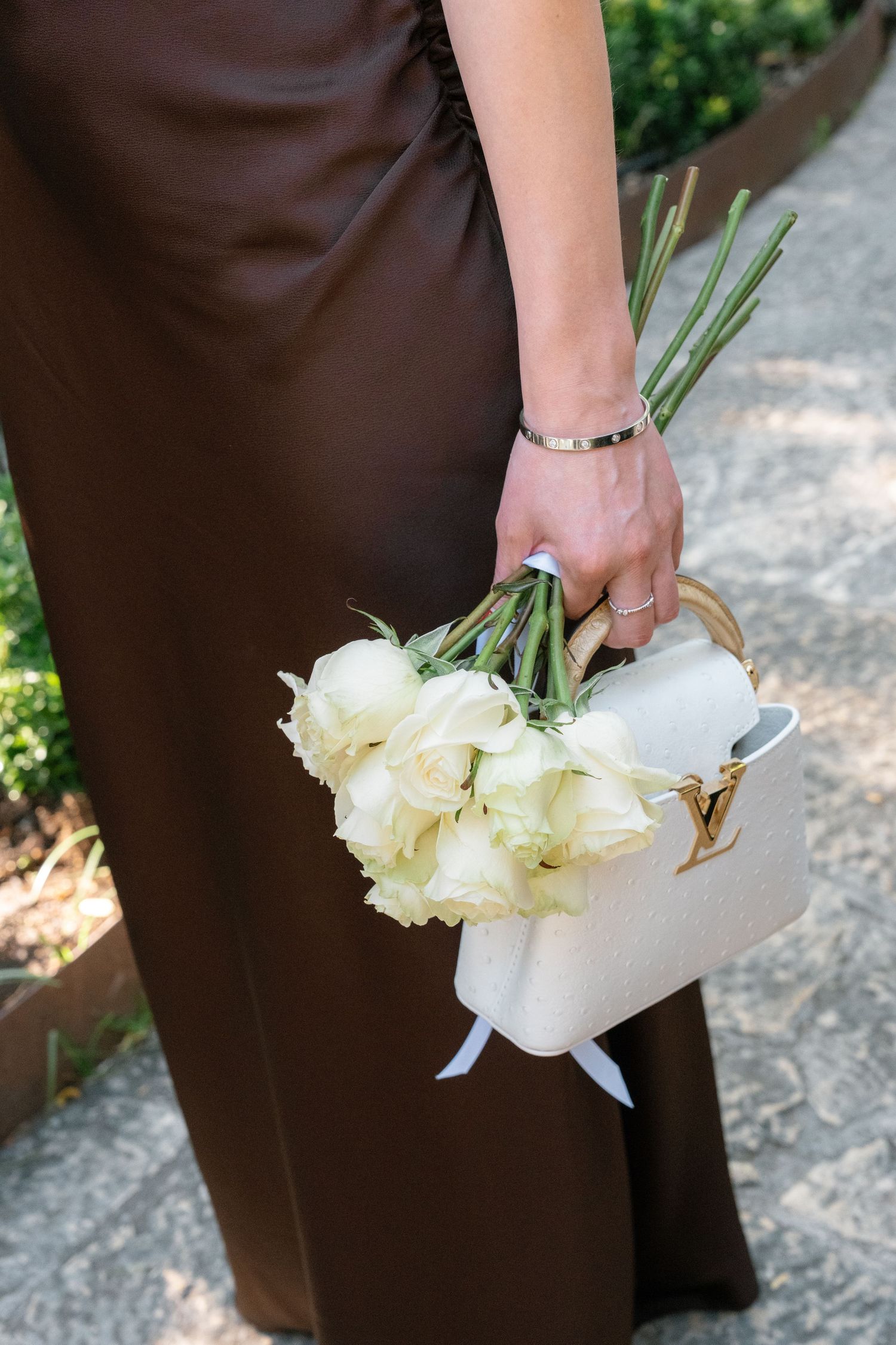 Woman in brown outfit holding white roses bouquet and a Louis Vuitton white handbag outdoors.