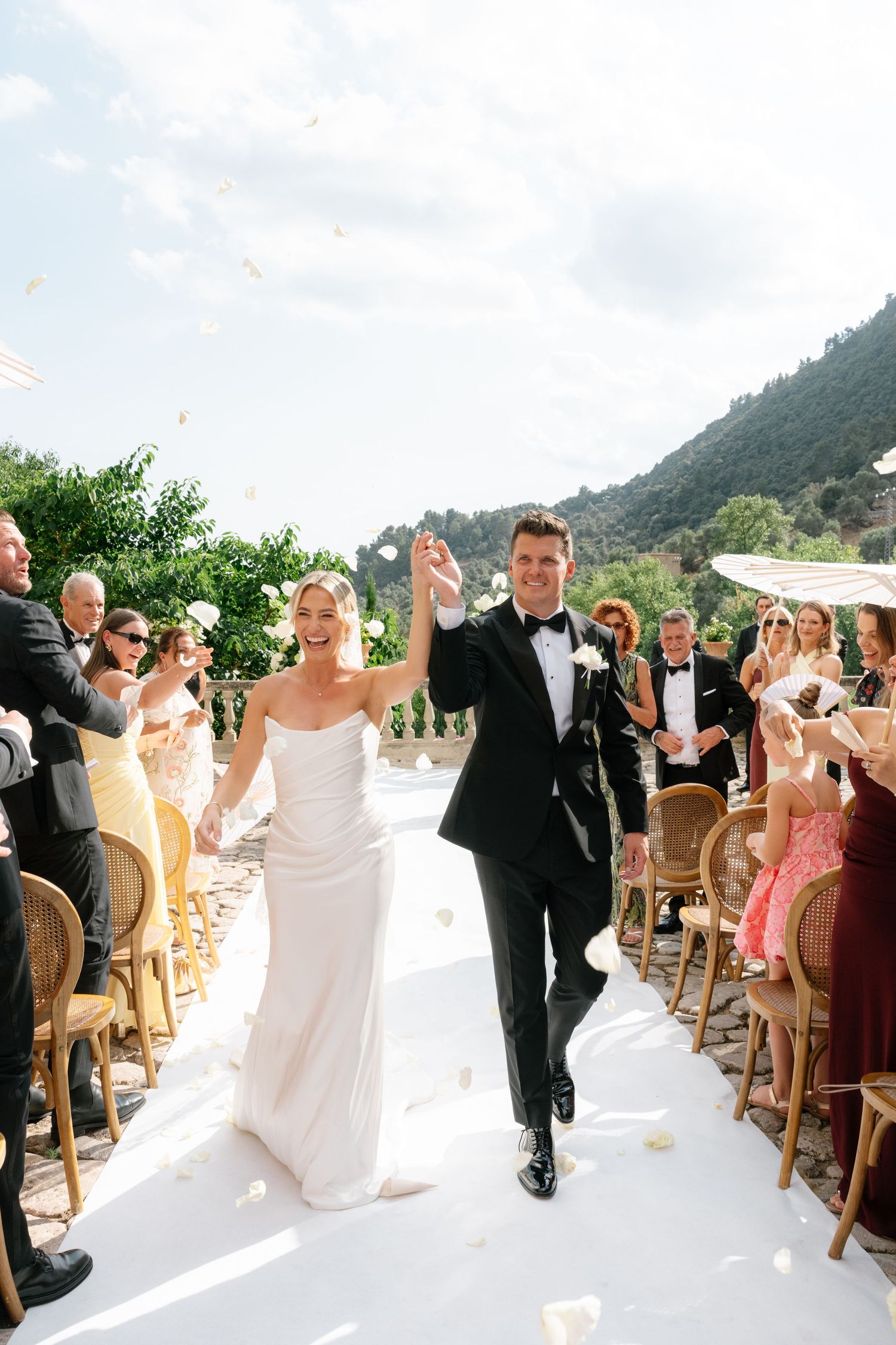 Joyful newlyweds walk down white aisle as guests toss petals at outdoor wedding ceremony with mountain backdrop.