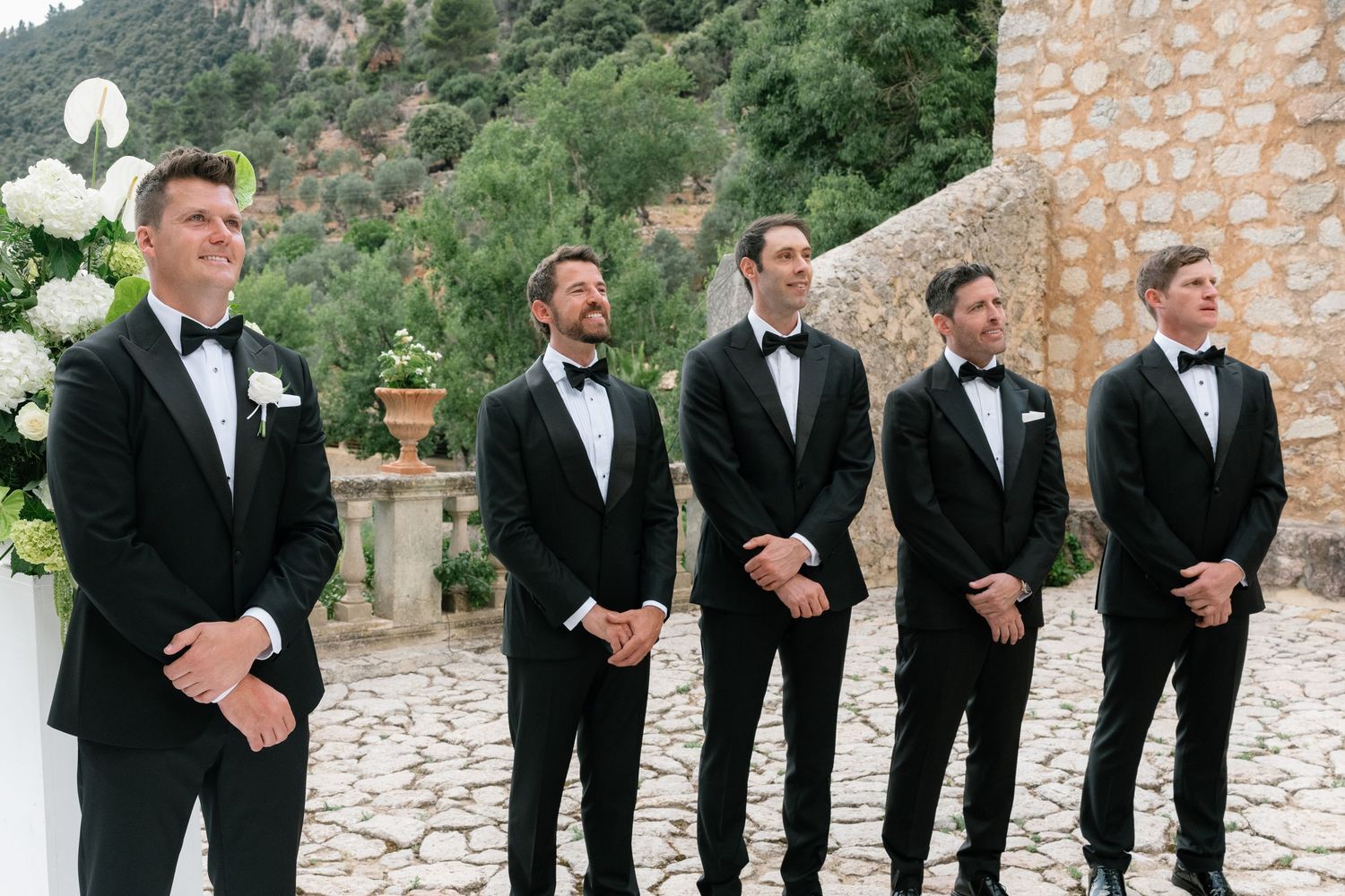 Five groomsmen in black tuxedos stand outdoors at a wedding ceremony with lush greenery and stone architecture.
