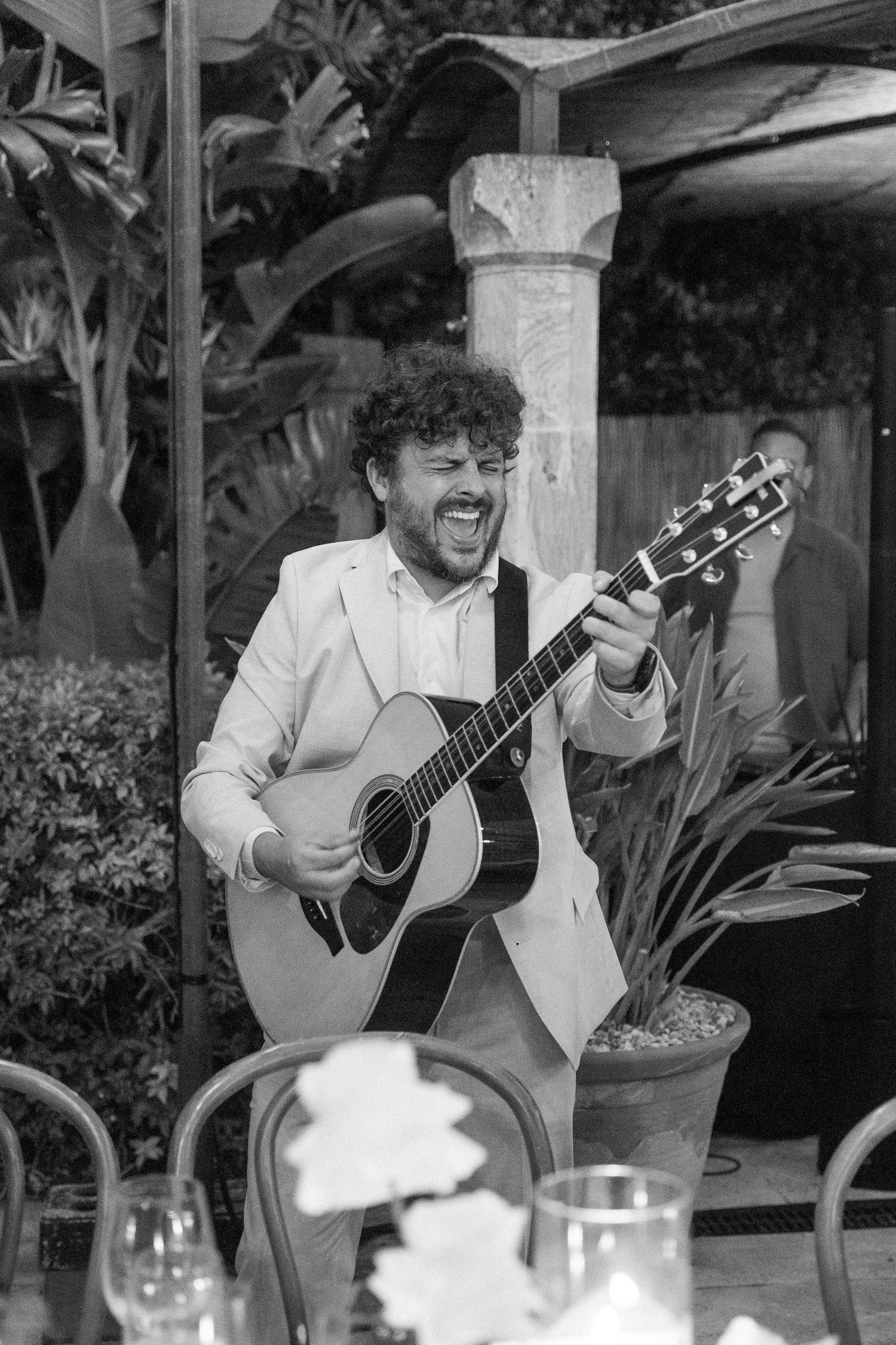 Musician in white suit passionately performs on acoustic guitar at outdoor wedding reception.