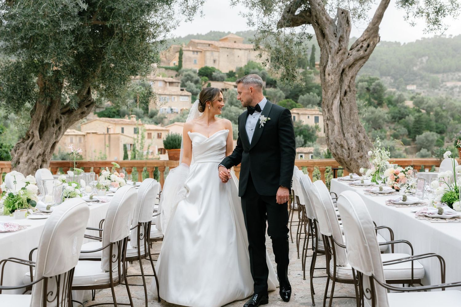 Outdoor wedding reception setup with white chairs and olive trees against Mediterranean backdrop.