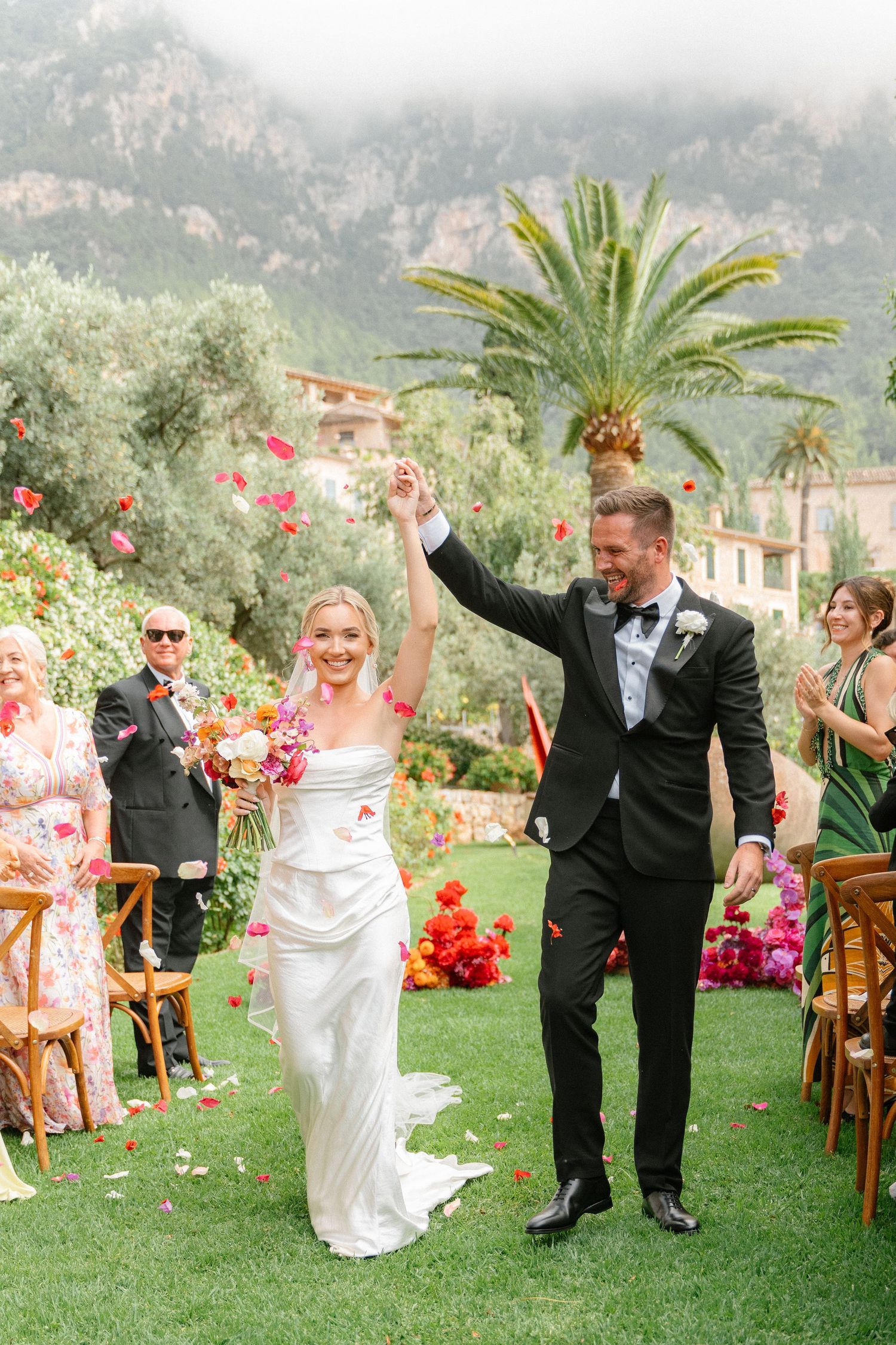 Joyful wedding couple celebrates with guests on a lush green lawn surrounded by tropical palm trees and pink flowers.