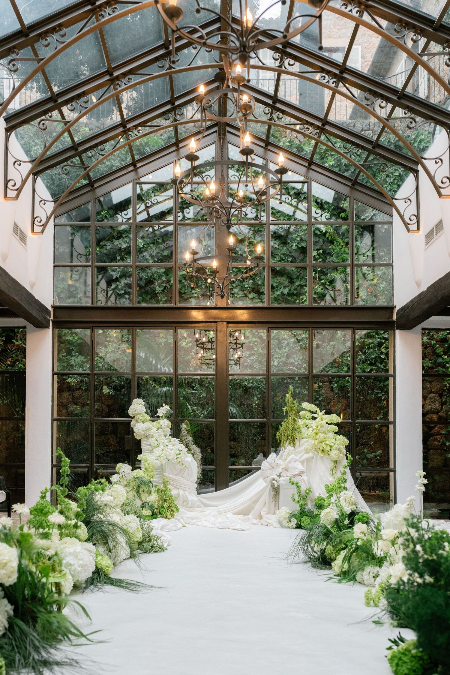Elegant greenhouse wedding venue decorated with white floral arrangements and hanging lights under glass ceiling.