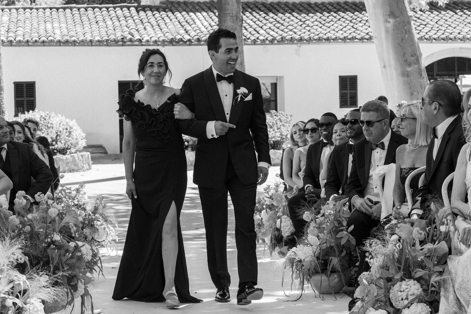 Black and white photo of a groom in a tuxedo escorting a woman in a gown at an outdoor wedding ceremony.