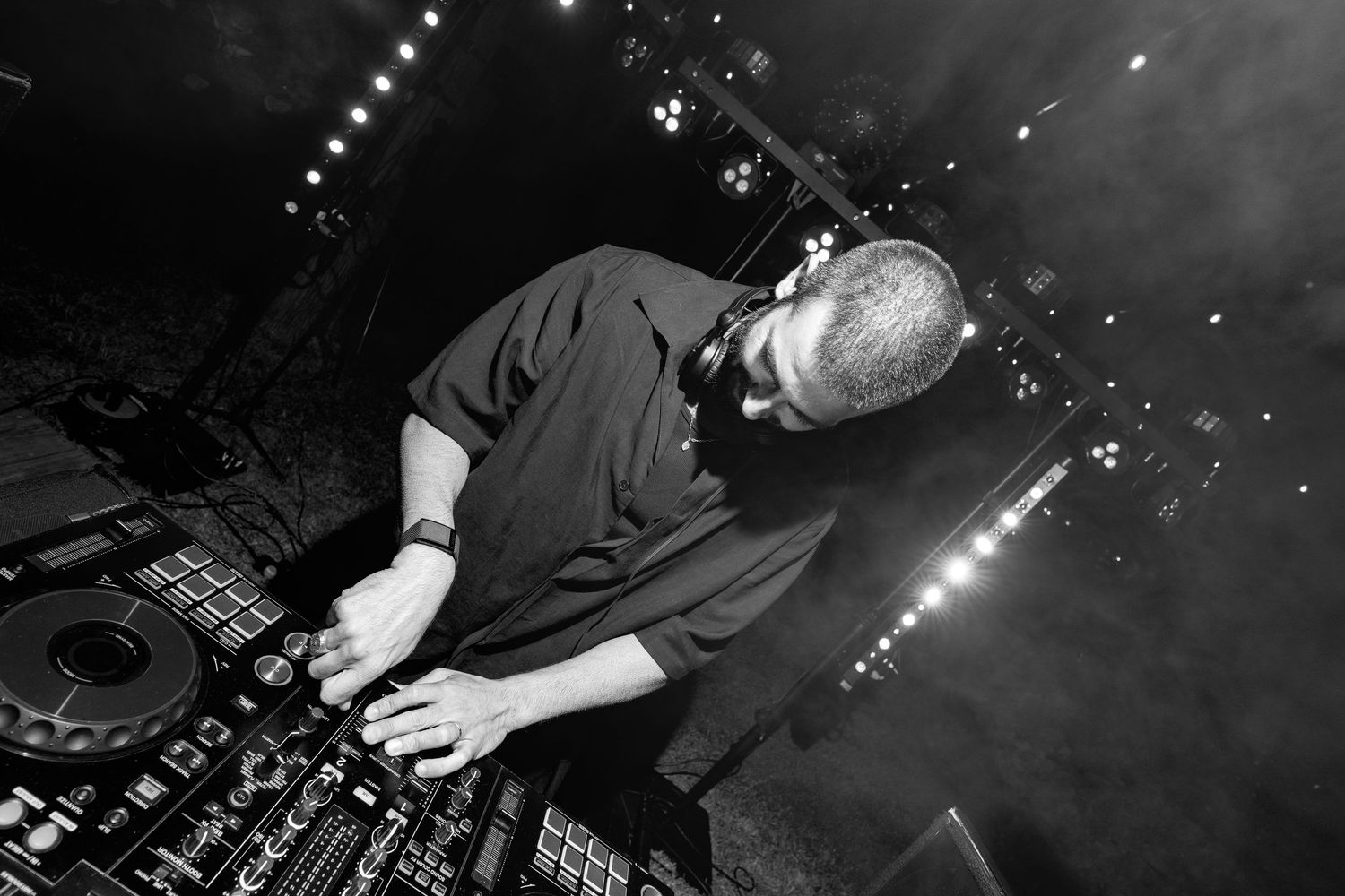 DJ performing at a nightclub, leaning over turntables under stage lights in black and white photo.