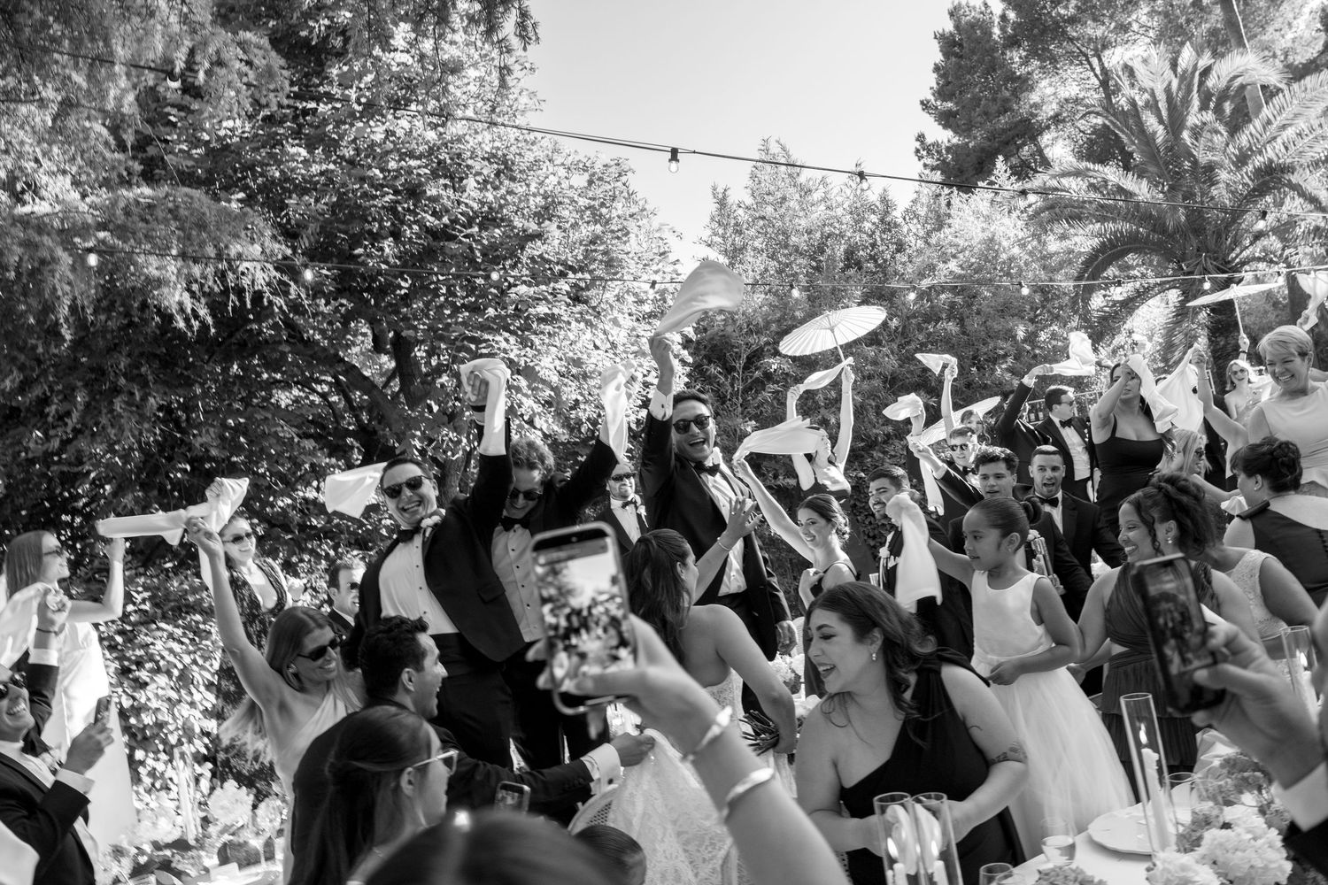 Black and white wedding photo of joyful guests waving white fans outdoors surrounded by lush trees.