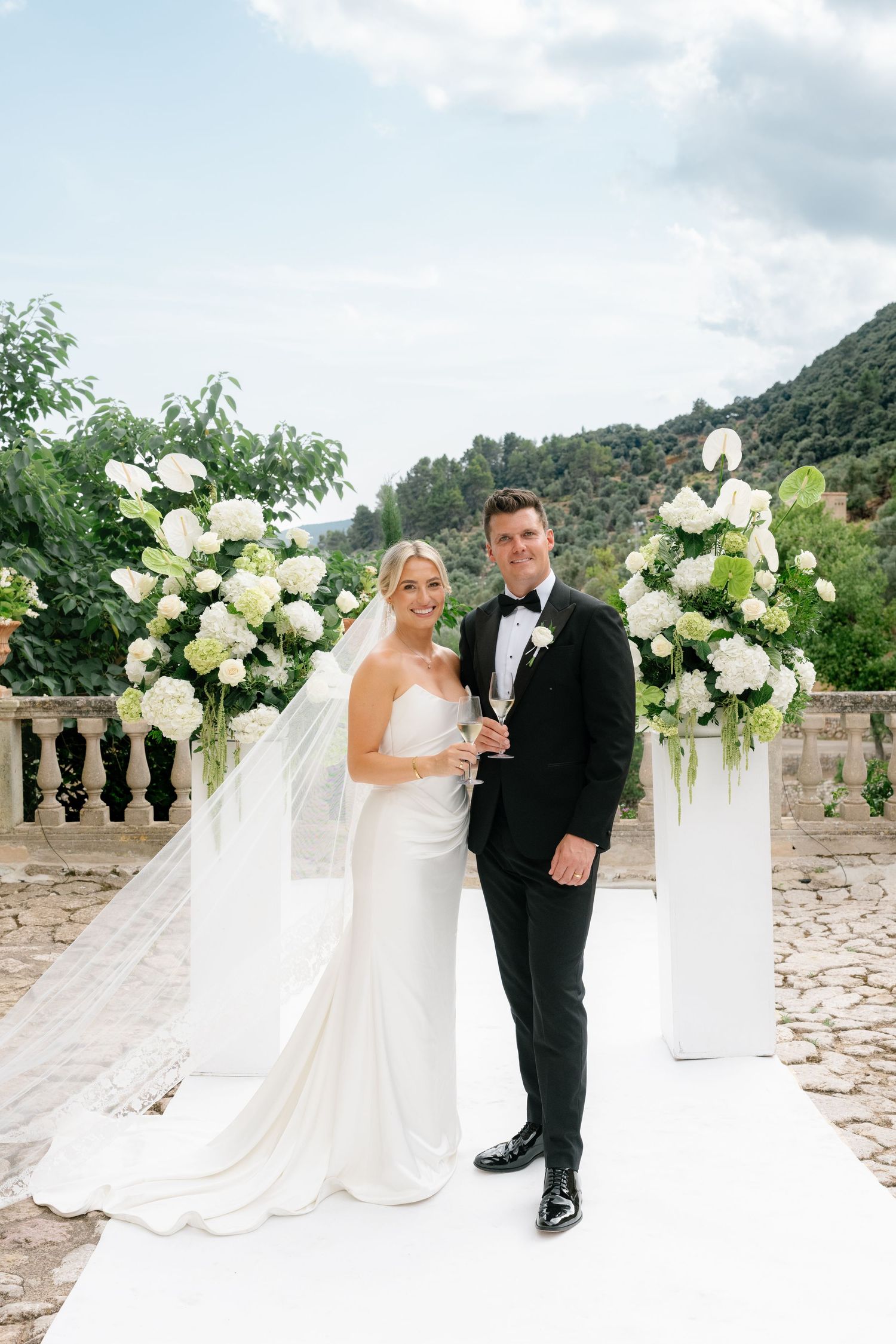 Bride and groom pose at outdoor wedding ceremony with white floral arrangements and mountain backdrop.