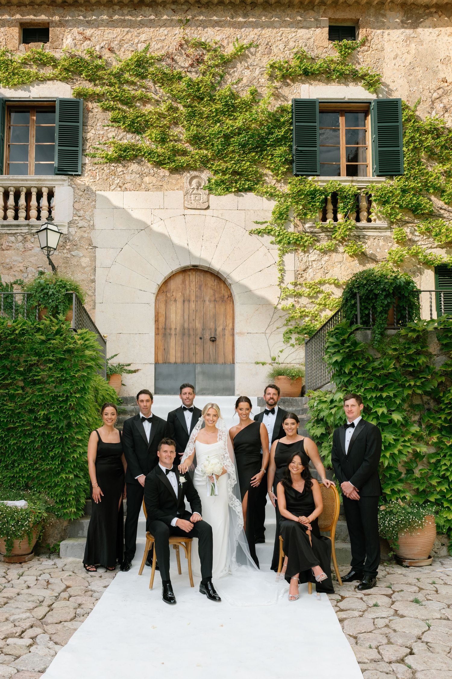 Wedding party in black attire poses before ivy-covered Italian villa with arched wooden door, bride in white gown centered.