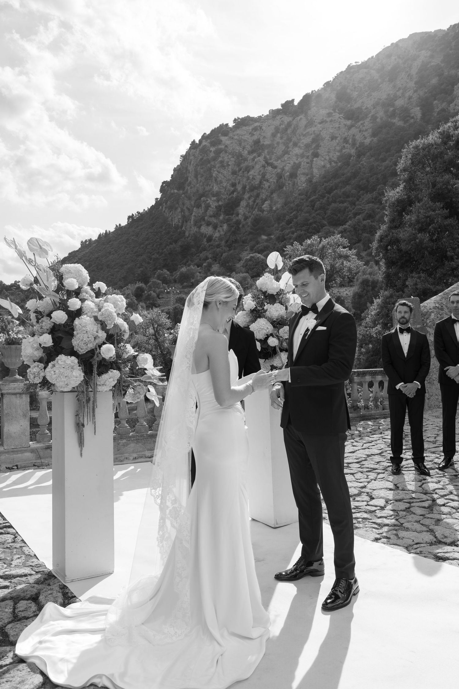 Black and white photo of bride and groom exchanging vows outdoors with floral arrangements and mountain backdrop.