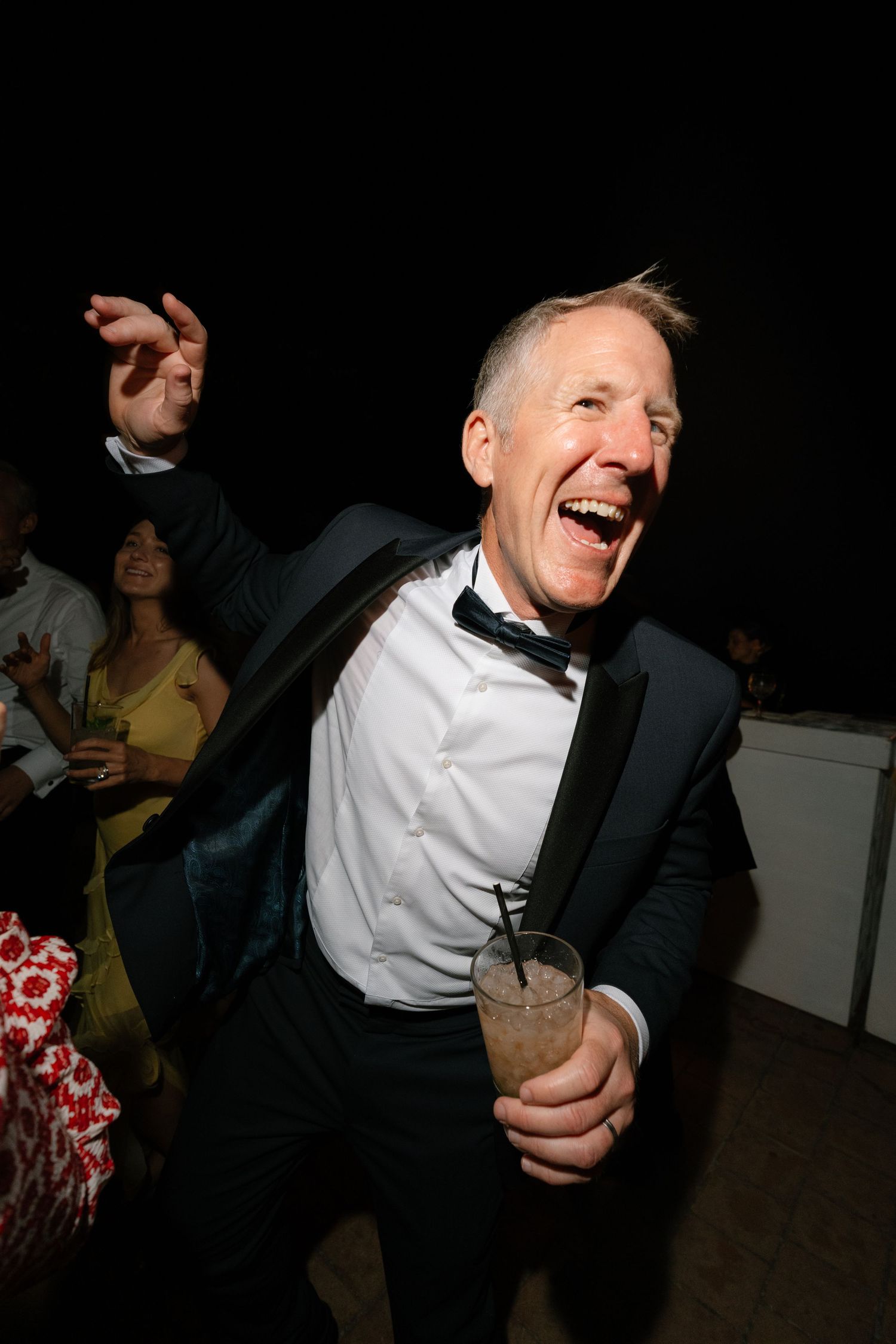 A joyful older man in a tuxedo laughs and dances while holding a cocktail at a formal evening event.