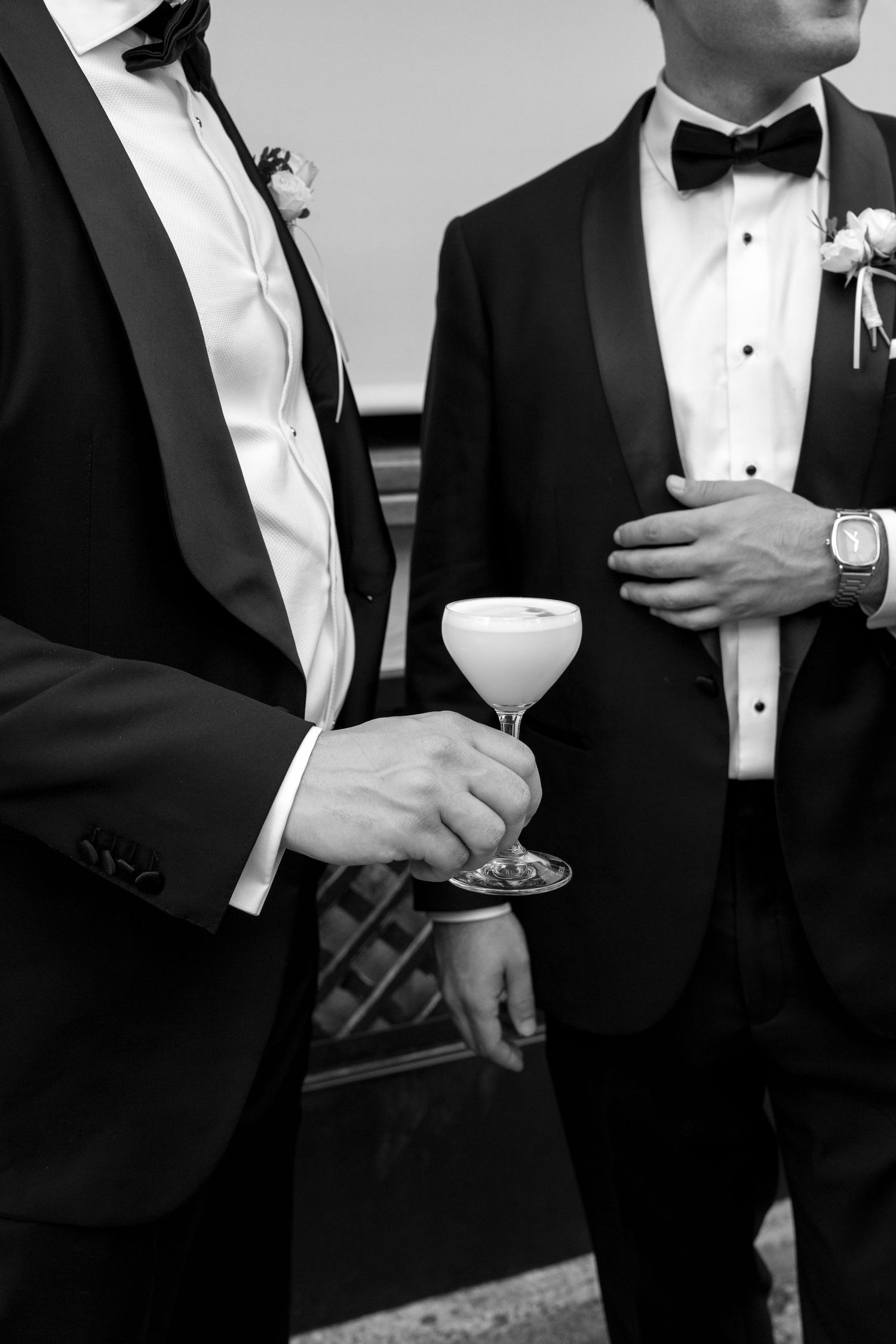 Two grooms in tuxedos with bow ties holding a cocktail glass at a wedding celebration.