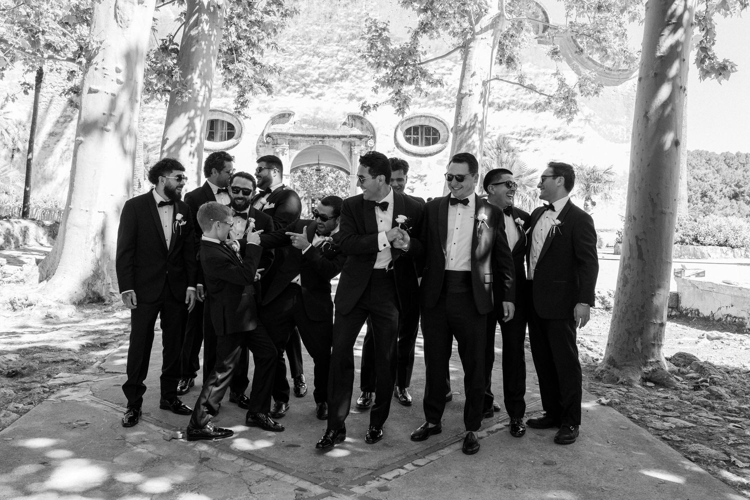 Black and white photo of groom and groomsmen in tuxedos laughing and walking together outdoors near vintage car.
