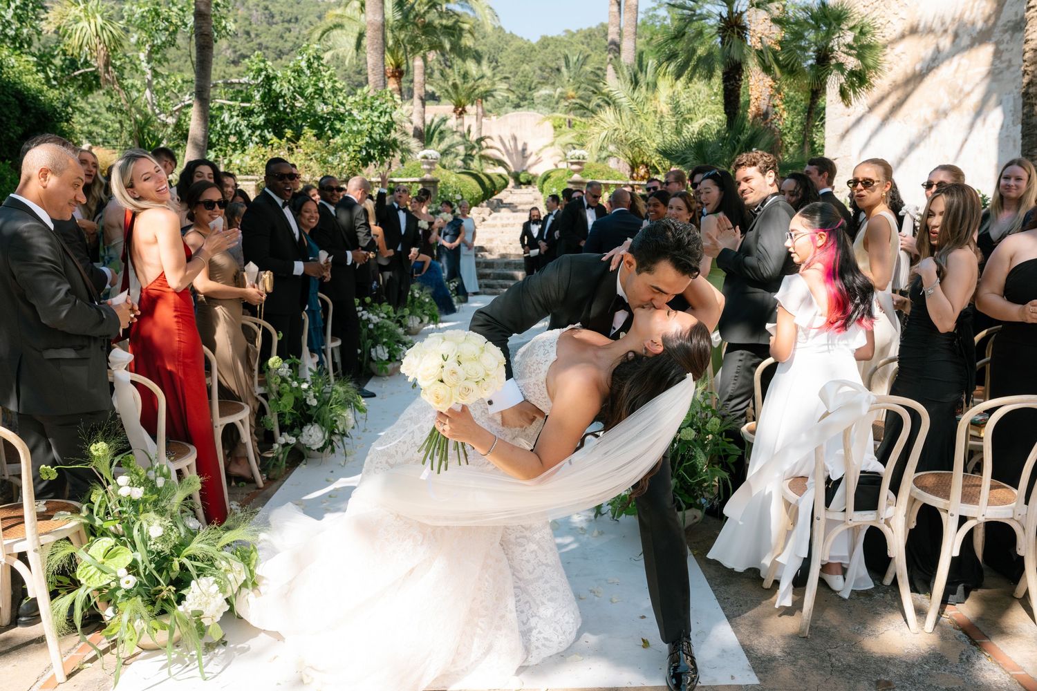 Groom dips bride for a romantic kiss while walking down the aisle at an outdoor wedding ceremony with guests cheering.