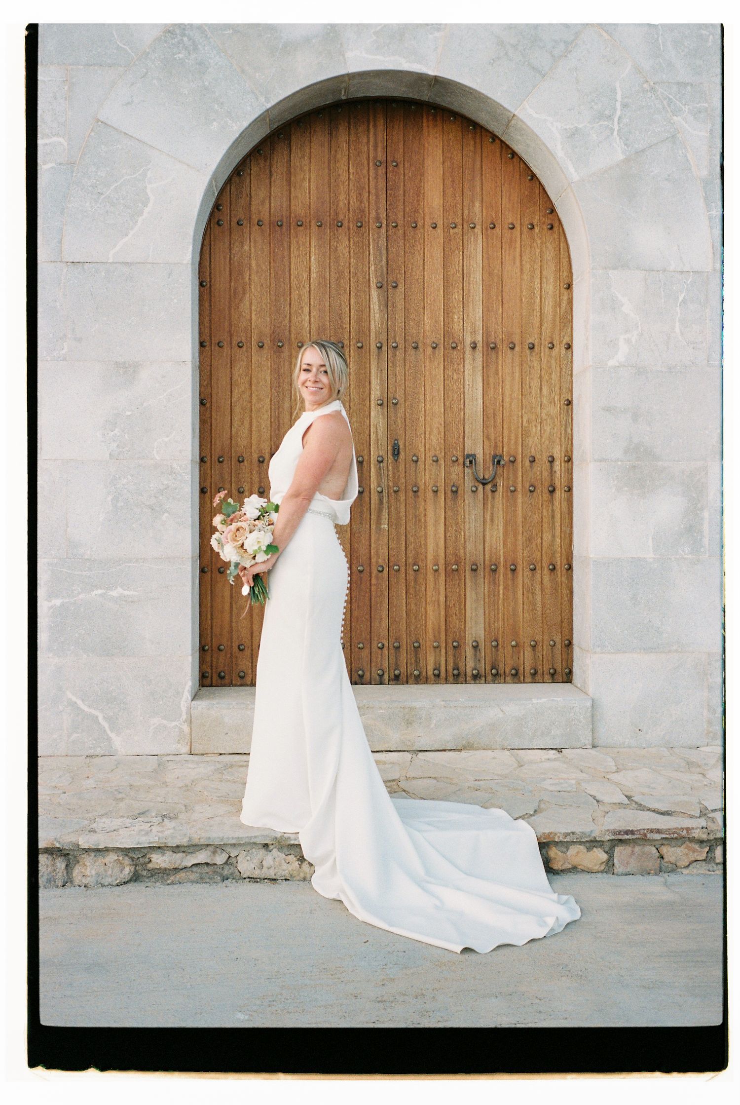 A bride in a flowing white dress poses against stone walls and wooden doors.