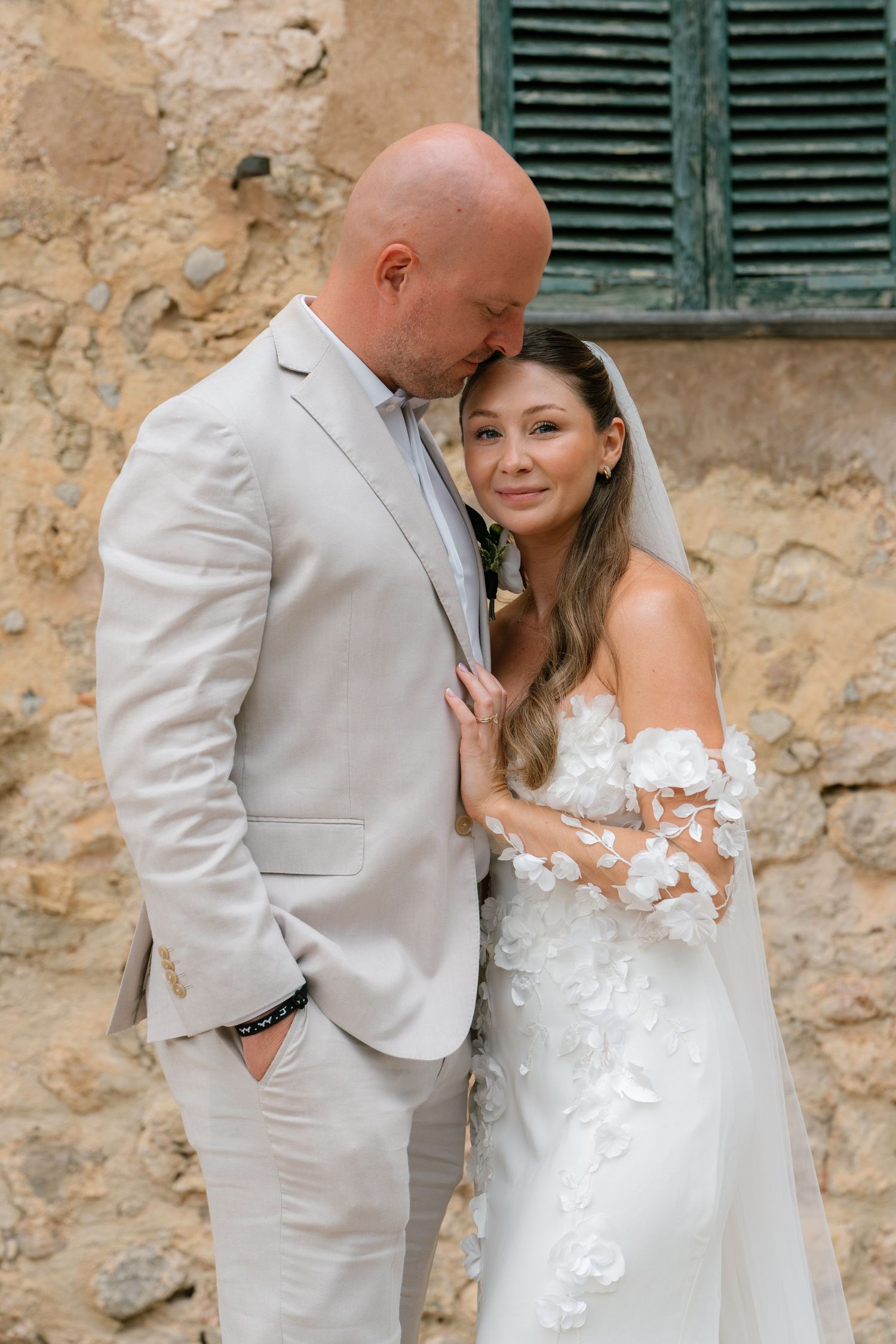 Wedding couple poses against rustic stone wall with green shutters.