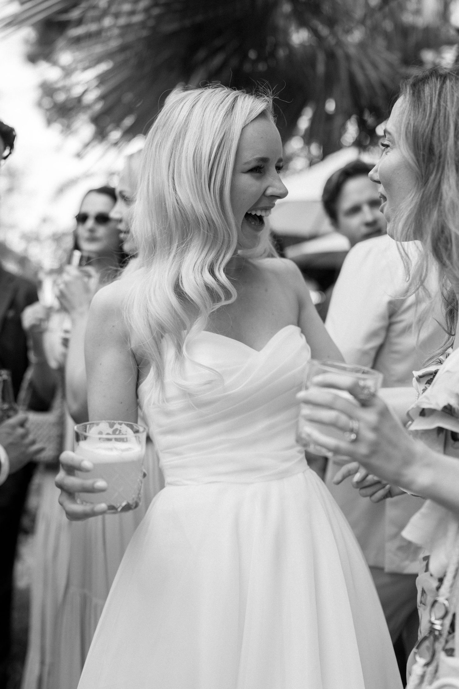 Wedding party members in white dresses enjoy drinks and conversation during an outdoor reception.
