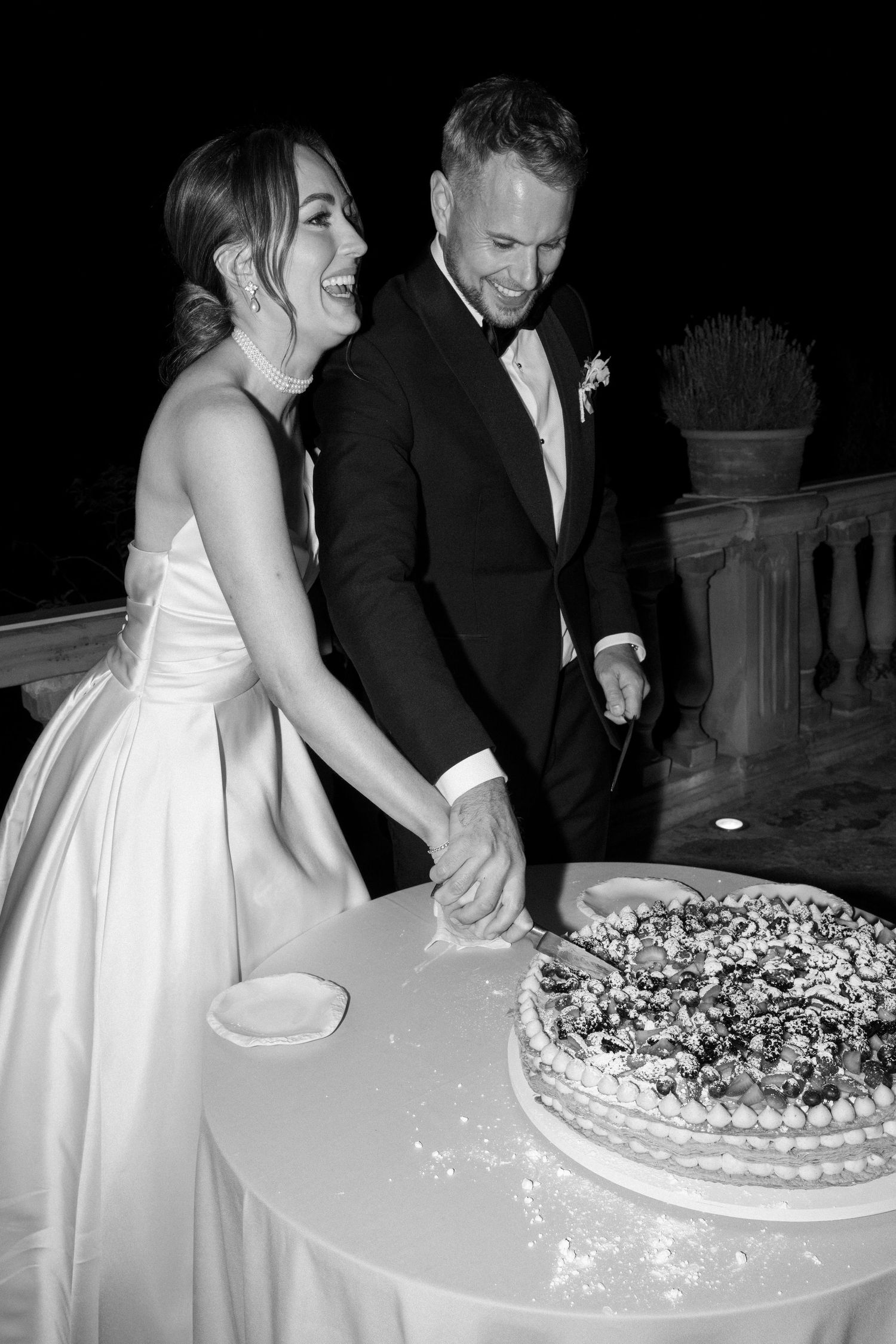 Black and white photo of couple sharing a joyful moment while cutting their wedding cake.