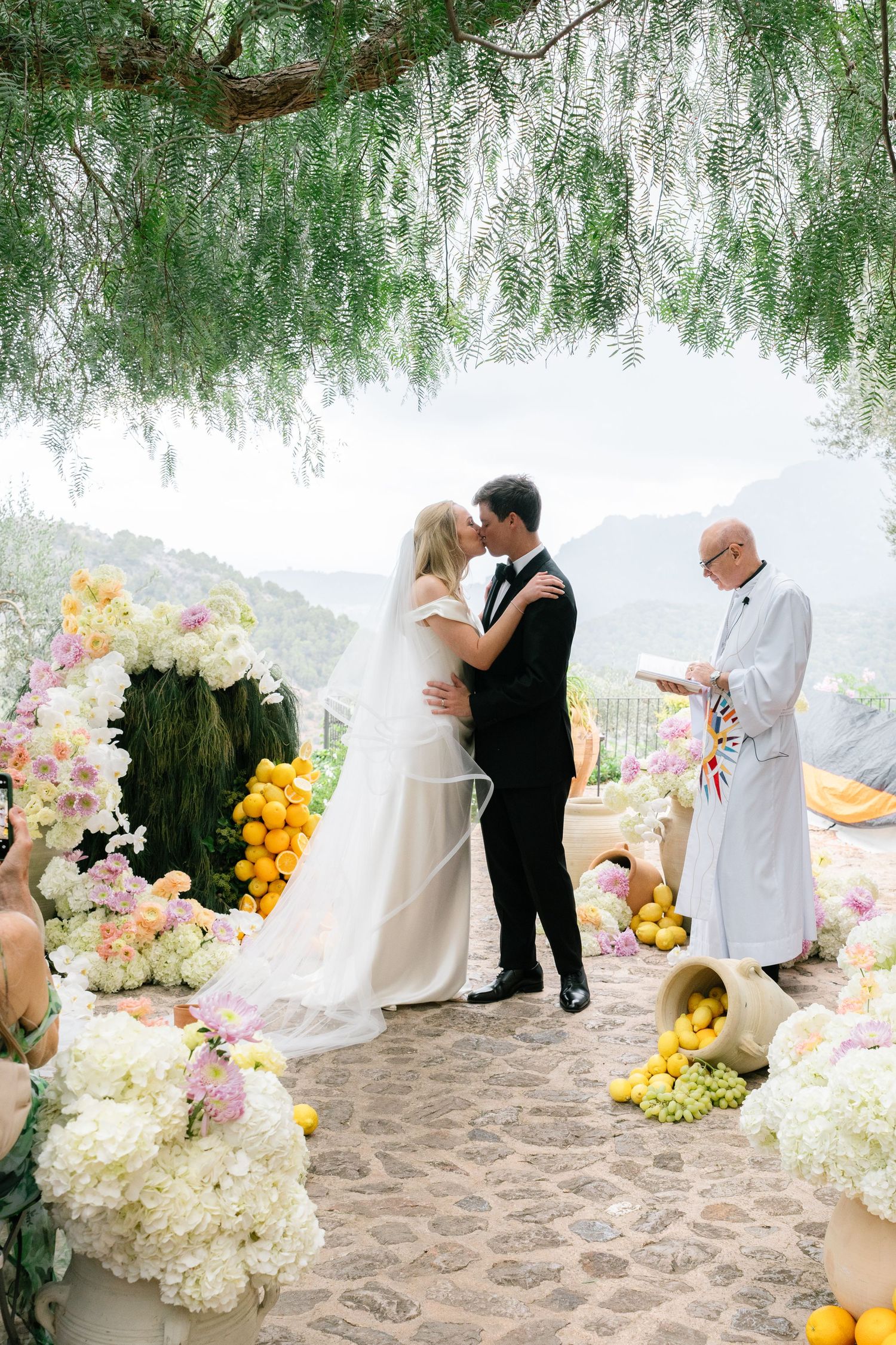 Wedding ceremony in progress under drooping willow branches with white and yellow floral decorations.
